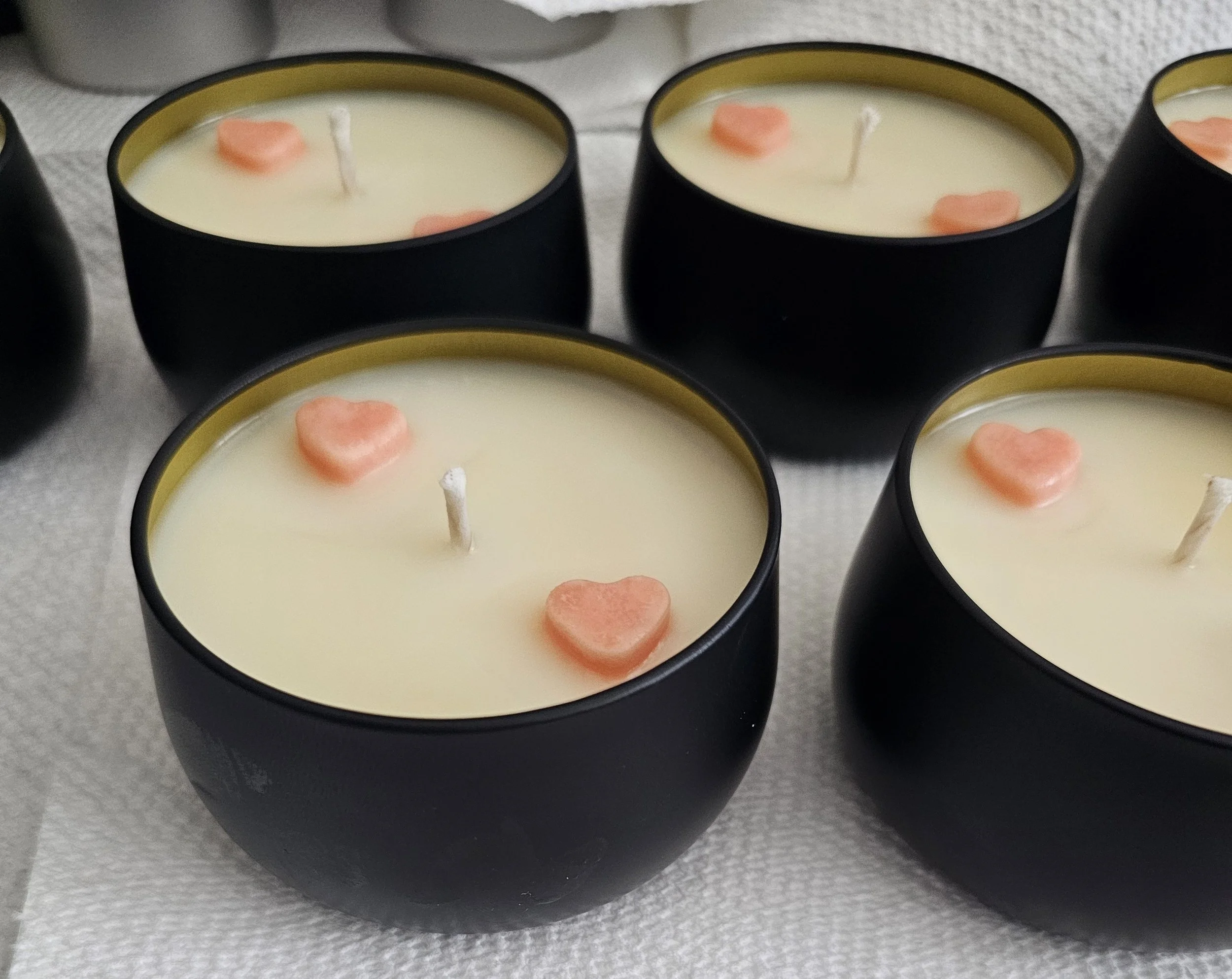 Several black candles with white wax, each decorated with a small pink heart-shaped object on top, are arranged on a gray surface.