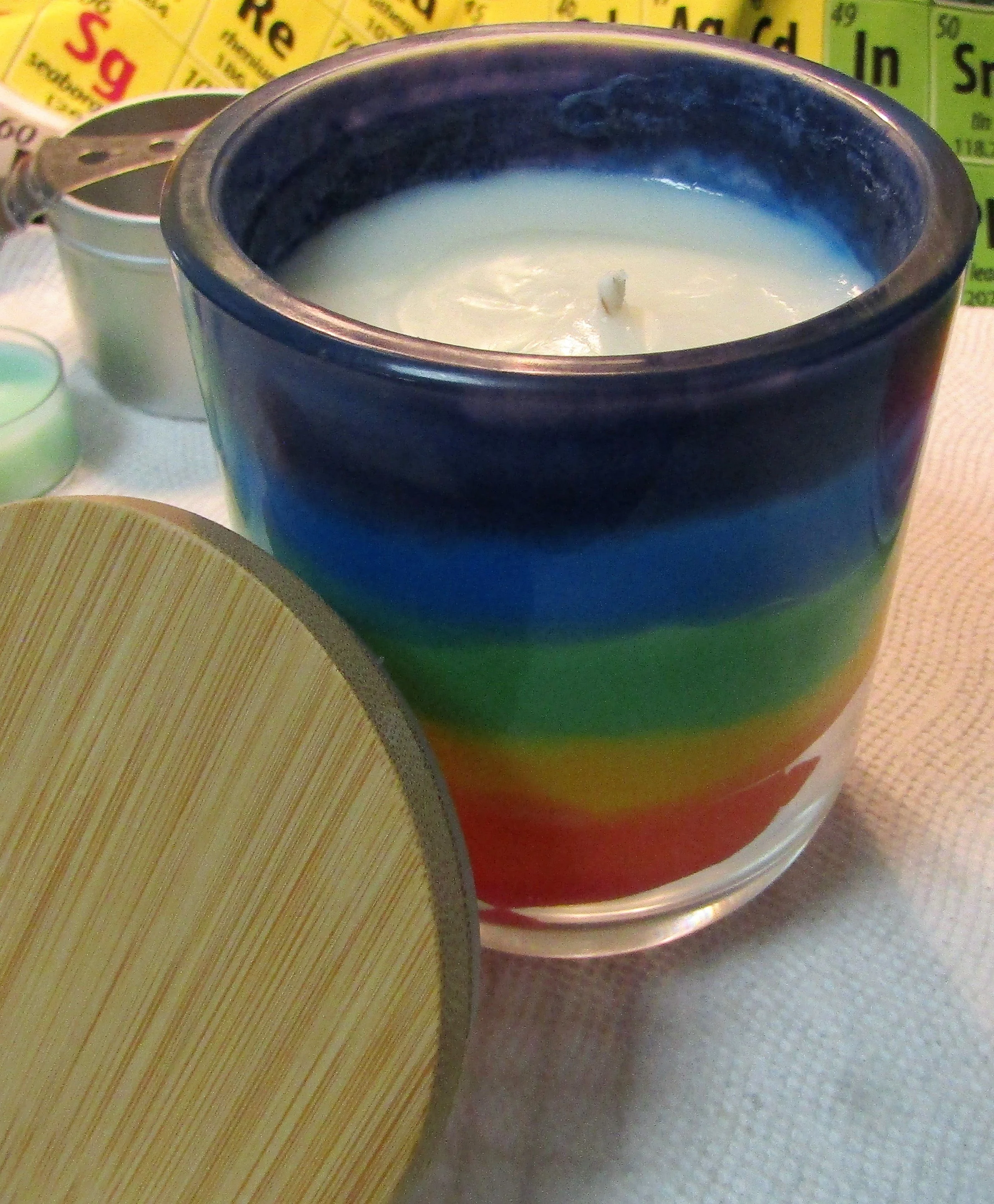 Close-up of a white candle in a black ceramic holder, placed on a wooden and paper placemat surface. In the background, there are colorful periodic table elements.