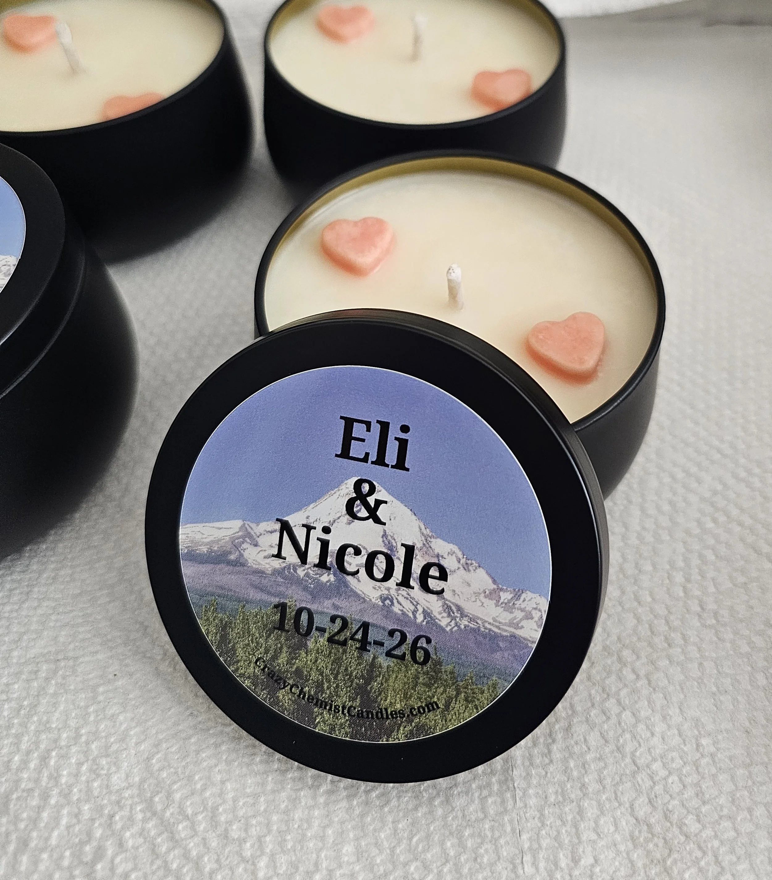 Close-up of three candles in black containers with pink heart-shaped wax pieces and unburned wicks, placed on a light-colored surface. One candle has a label with a mountain landscape and the text 'Eli & Nicole' and a date below.