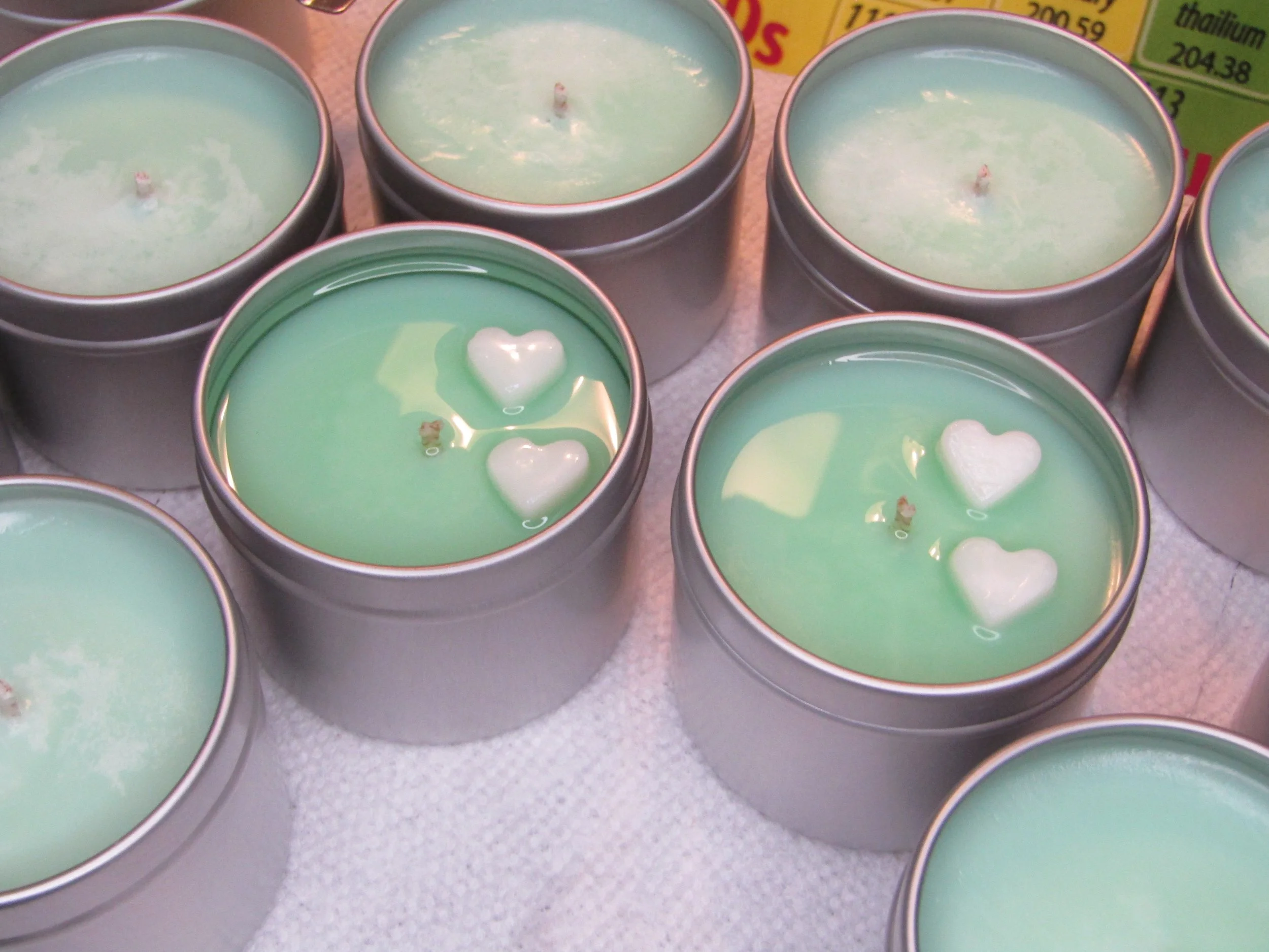 Multiple small candles in round metal tins with pale green wax, some with white heart-shaped decorations on top, placed on a white surface.