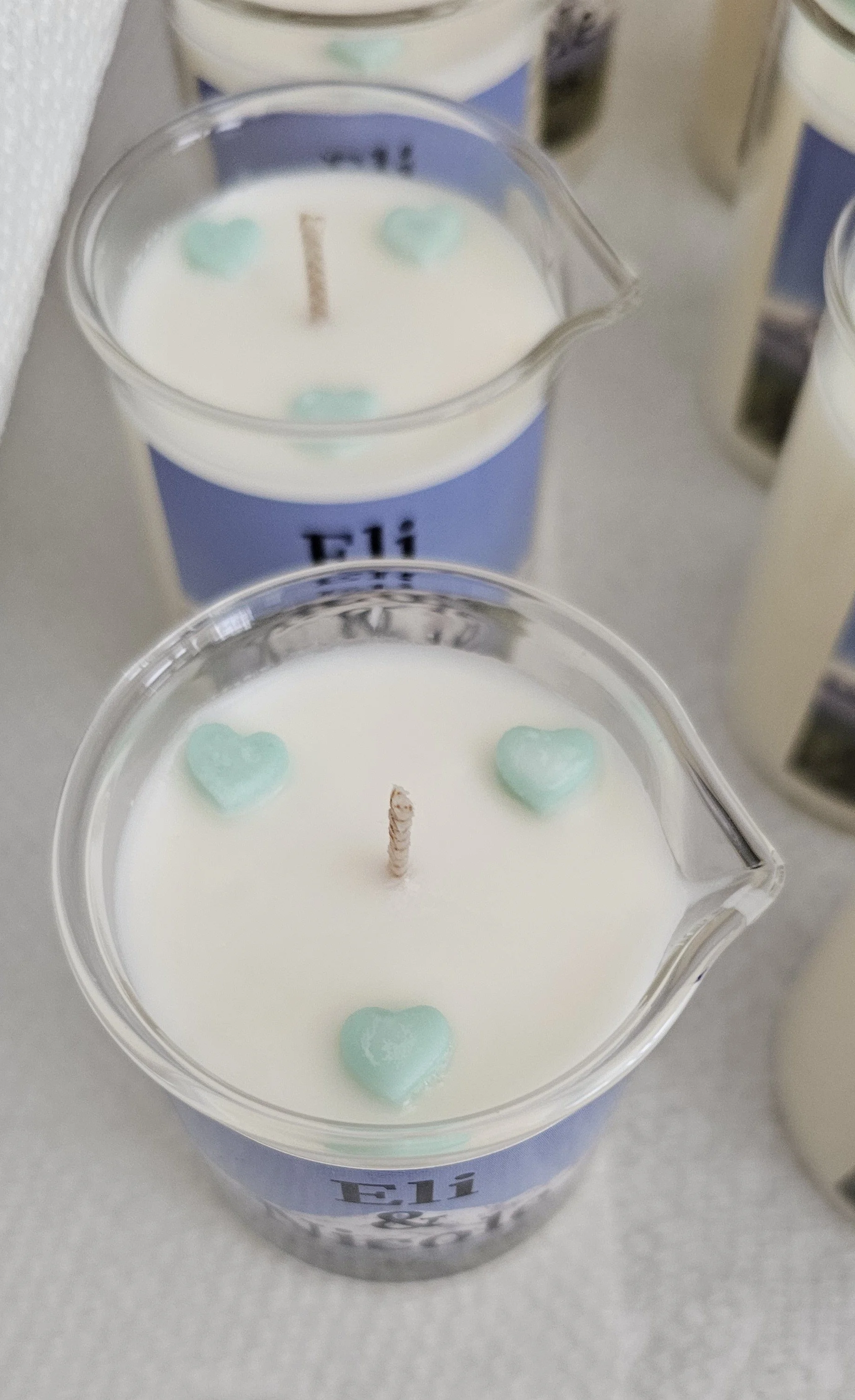 A white candle in a glass holder decorated with light blue heart-shaped ornaments and a single wick in the center, placed on a flat surface with similar candles in the background.