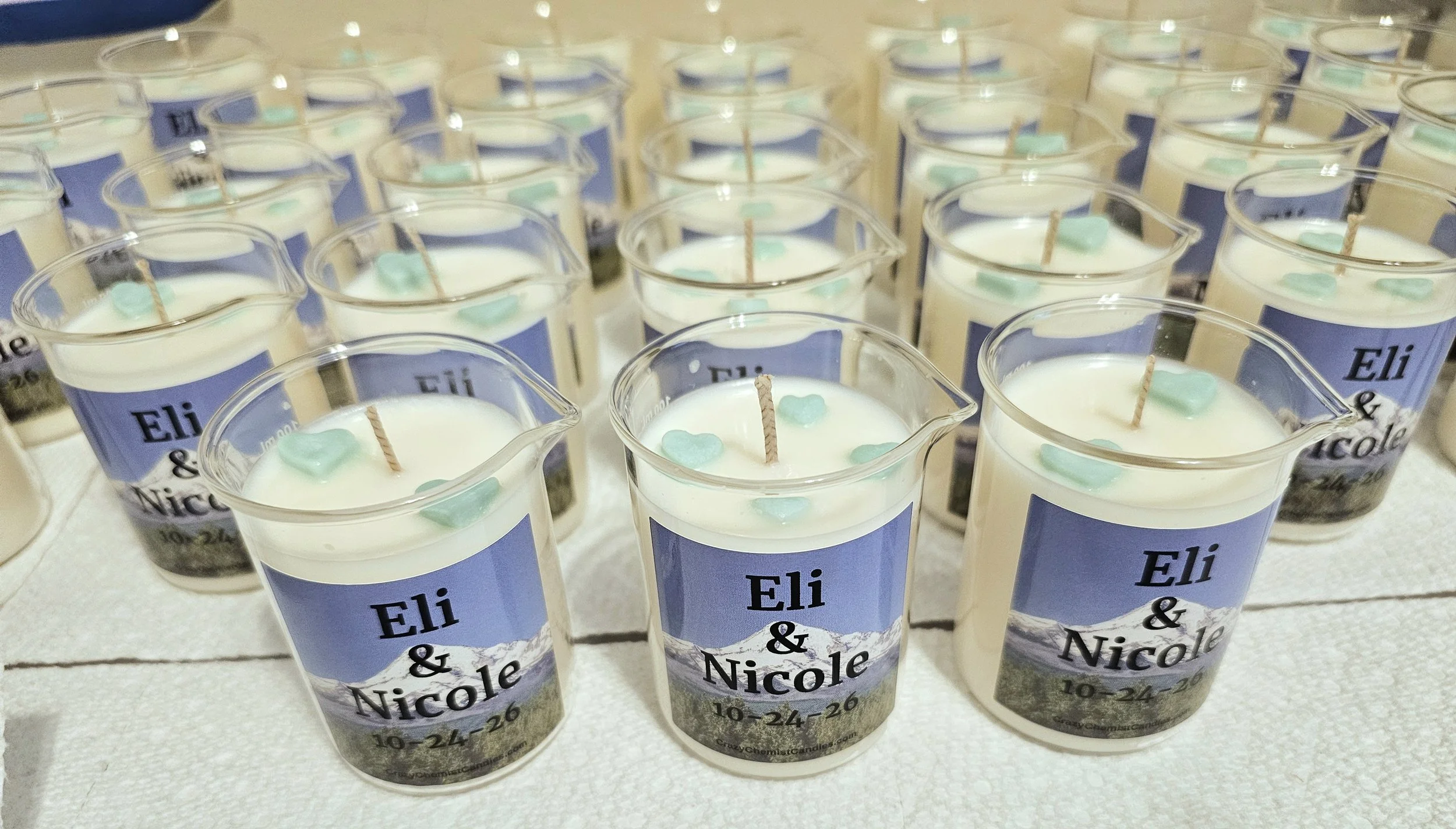 Multiple personalized candles in clear plastic cups with a mountain landscape label, each with a small heart-shaped decoration and a wick, likely for a wedding or event.