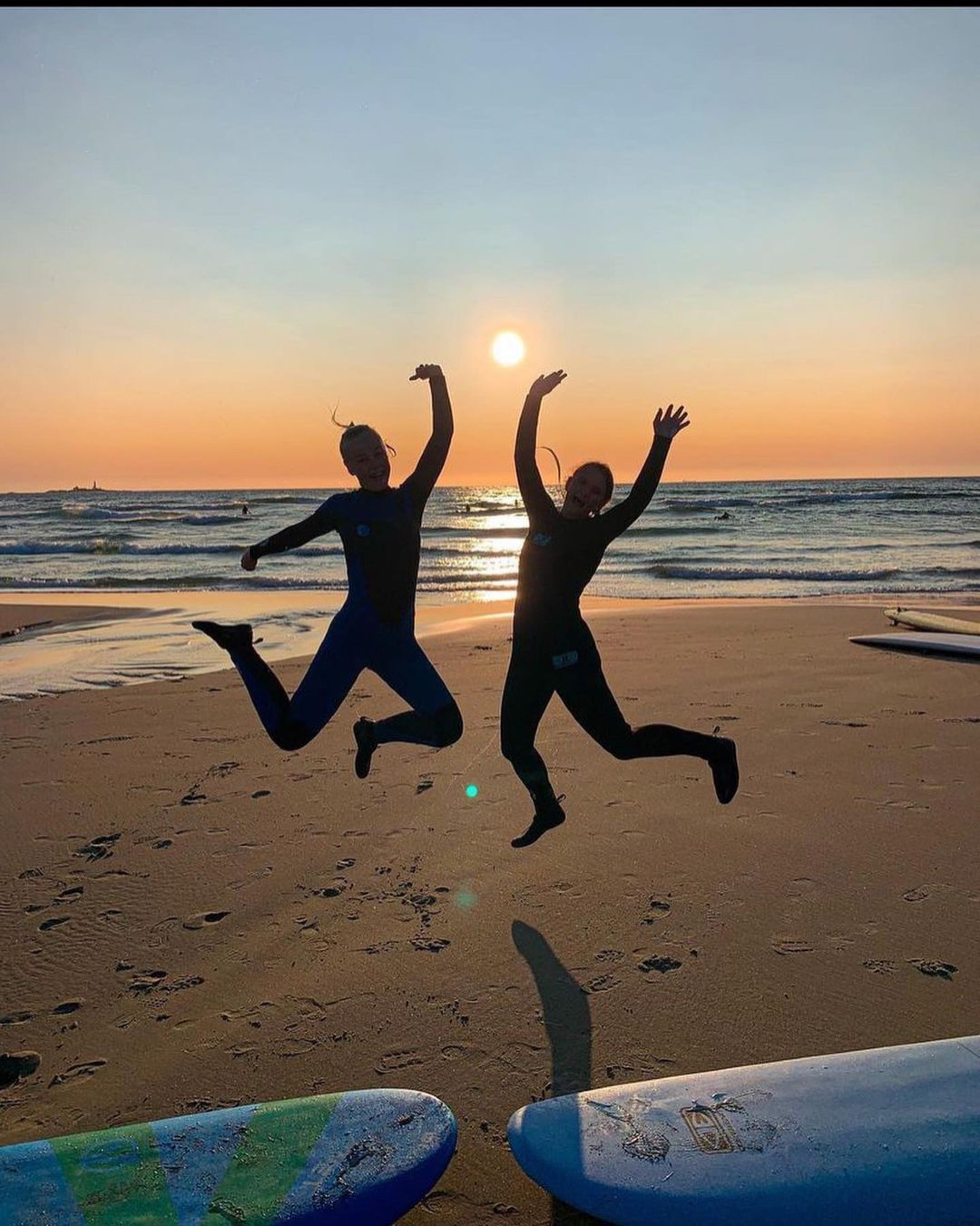 siluetter av to elever i surfedrakt som hopper på stranda i solnedgang.