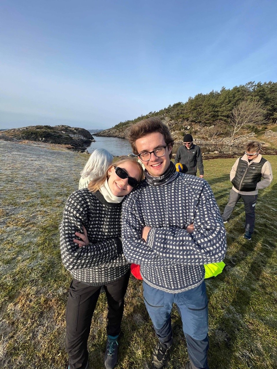 A boy and a girl from different countries in traditional patterned sweaters gathered on a grassy coastal landscape near the sea on a clear day.