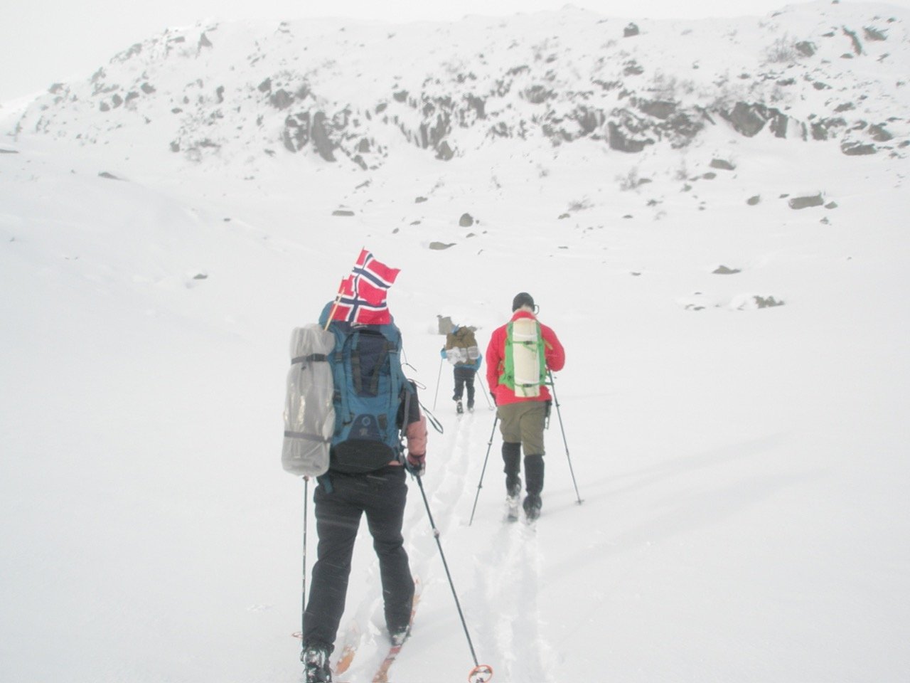 Three backcountry skiers carrying large backpacks travel through a snowy mountain landscape, with a Norwegian flag attached to one backpack