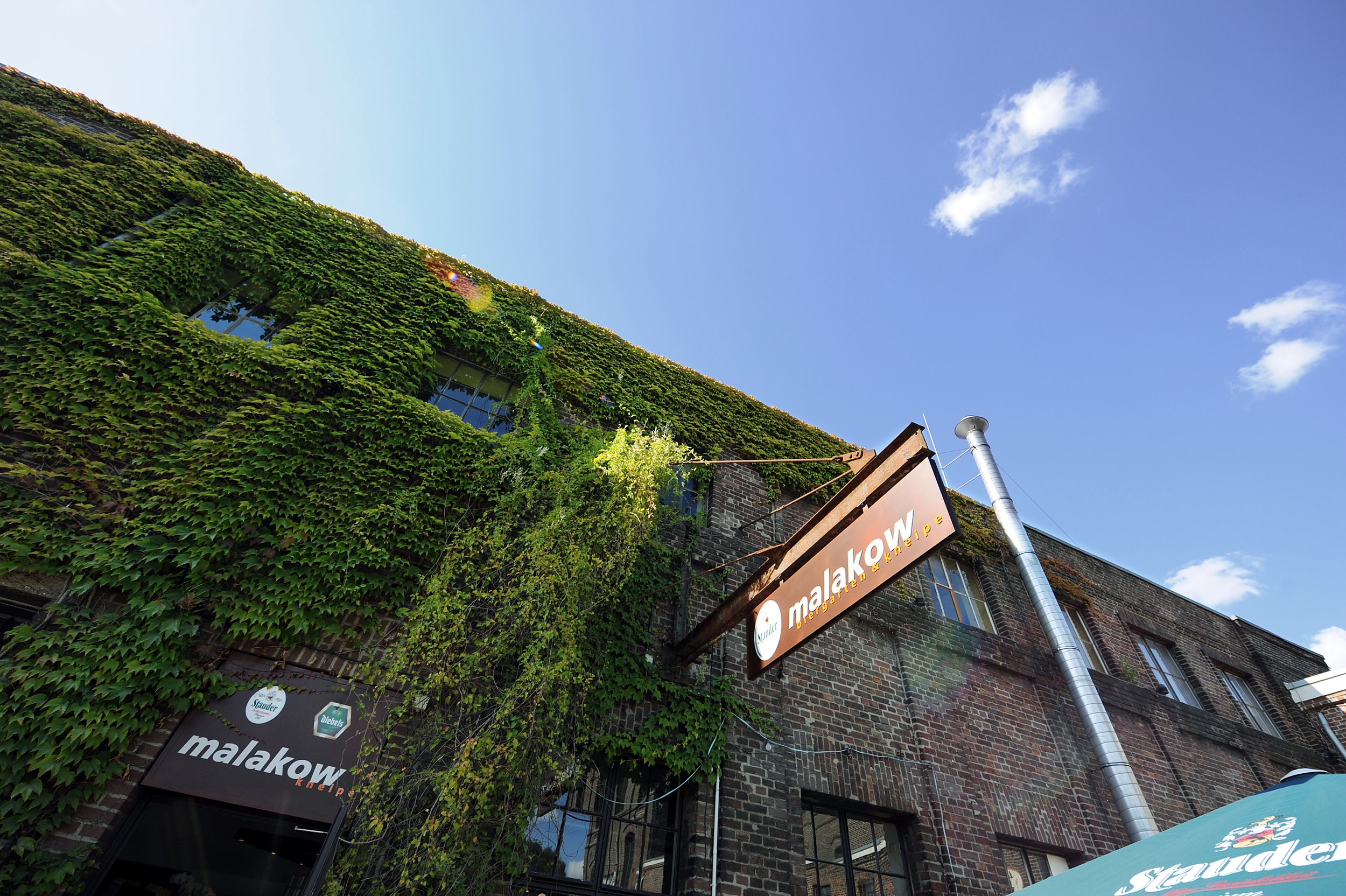Ein altes Fabrikgebäude mit Vollpflanzen an den Wänden, ein Schild mit der Aufschrift 'Malakow Kneipe', blauer Himmel mit wenigen Wolken.