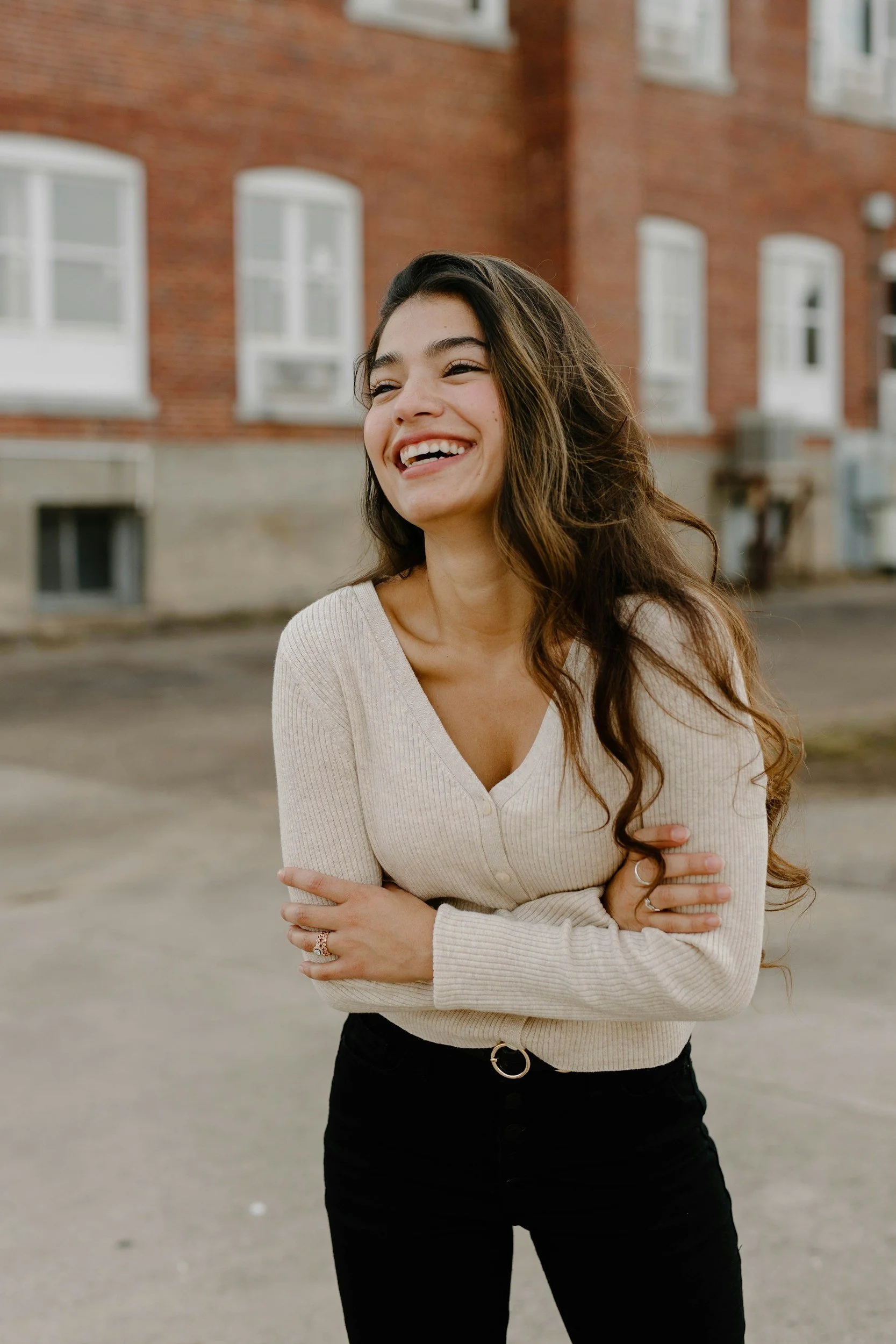 A young woman with long, wavy dark hair, smiling and laughing outdoors, wearing a beige ribbed sweater and black pants, standing in front of a brick building.