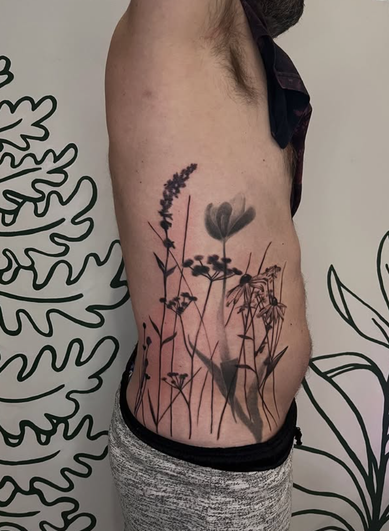 A person's side torso with a black and gray floral tattoo of wildflowers and plants on the ribcage area.