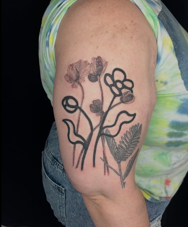 A tattoo of various flowers on a person's upper arm, combining black line art and shaded floral designs.