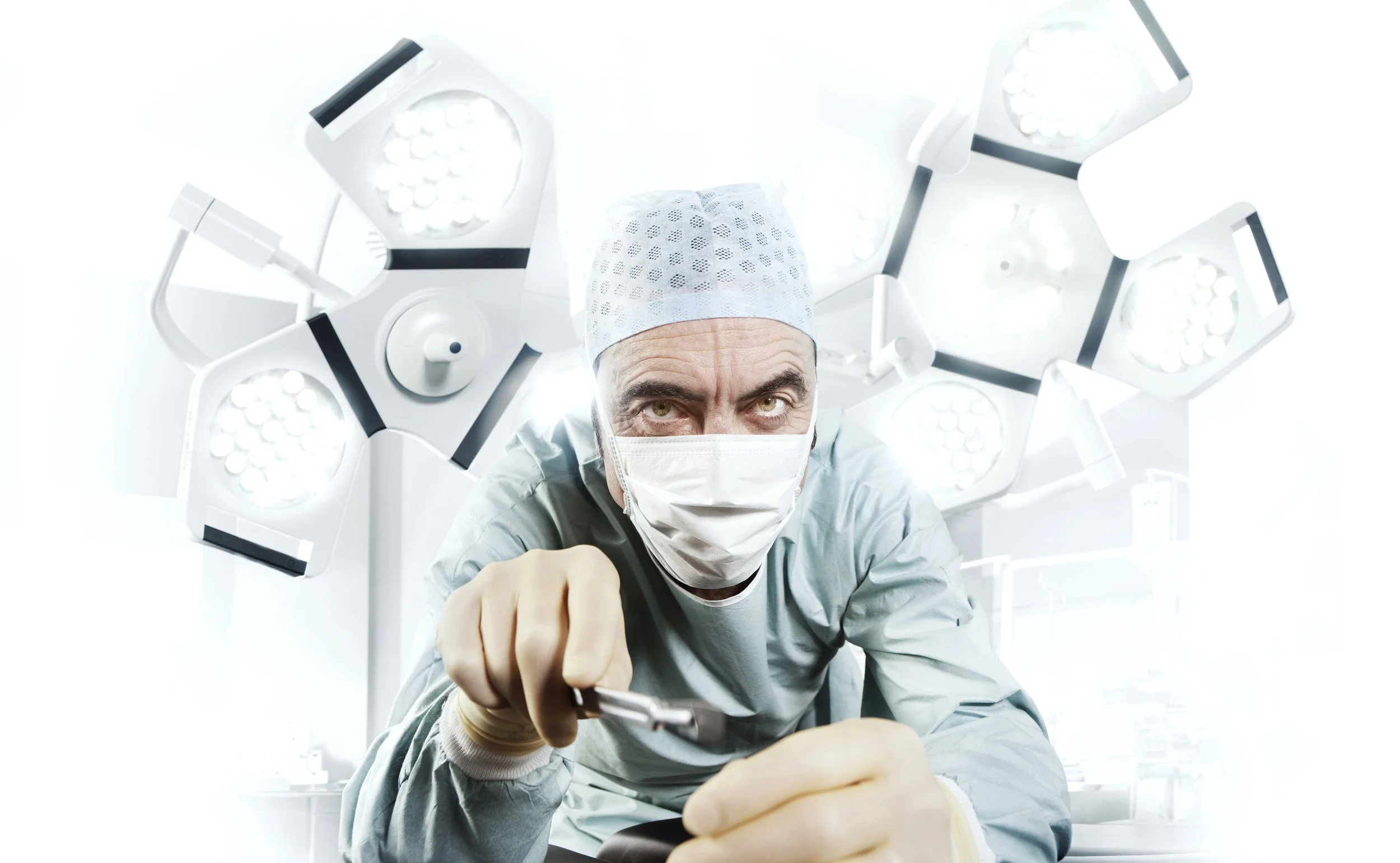 Promo image for the ITV show Monroe. James Nesbitt, dressed as a surgeon and holding a scalpel, leans in to camera in front of big surgery lights. entertainment portrait photography by Steve Bright, London