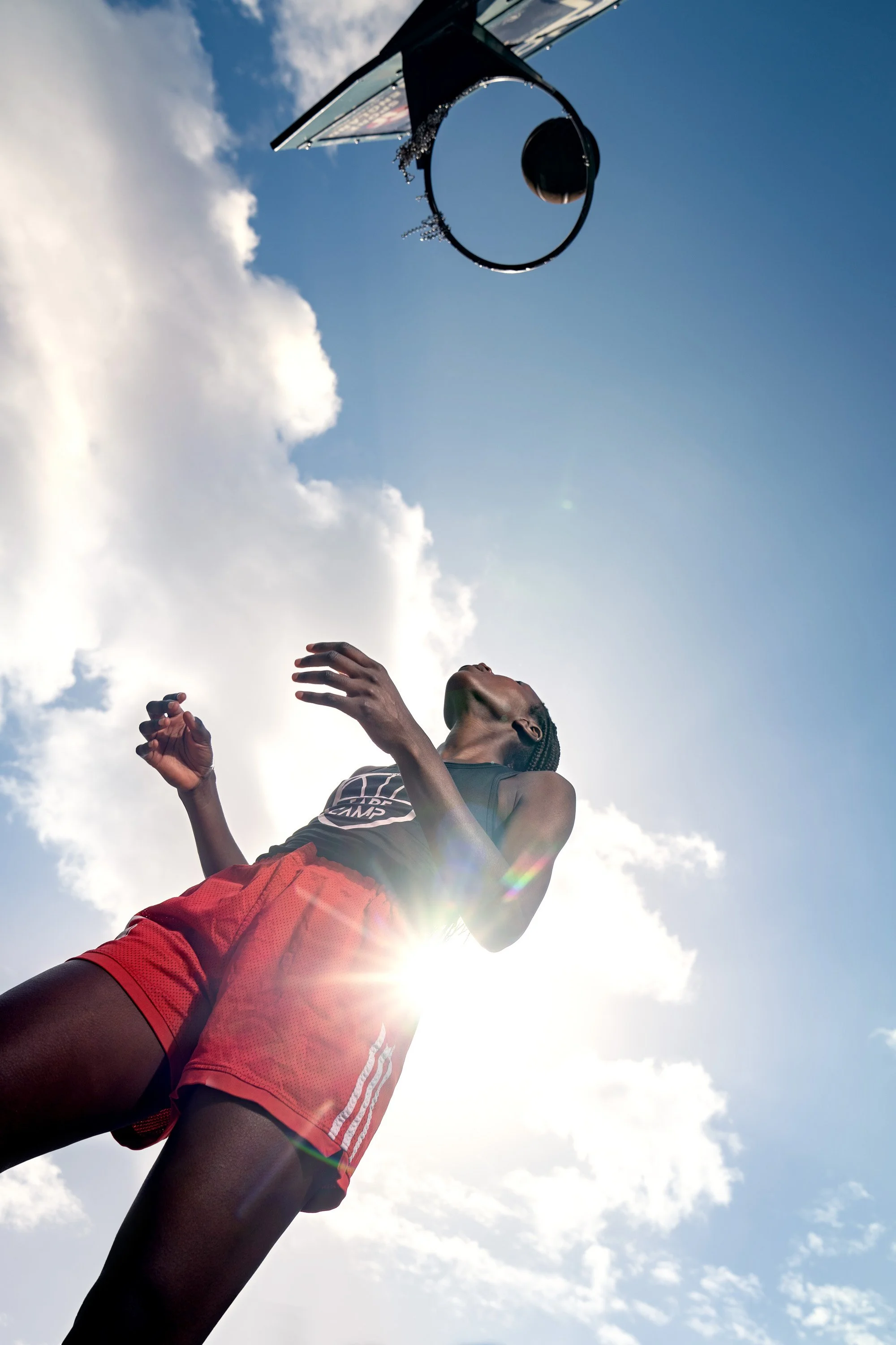 A Black female basketball player wearing red athletic shorts and a black top is playing basketball outdoors under a bright blue sky with clouds. dynamic sports portrait photography by Steve Bright