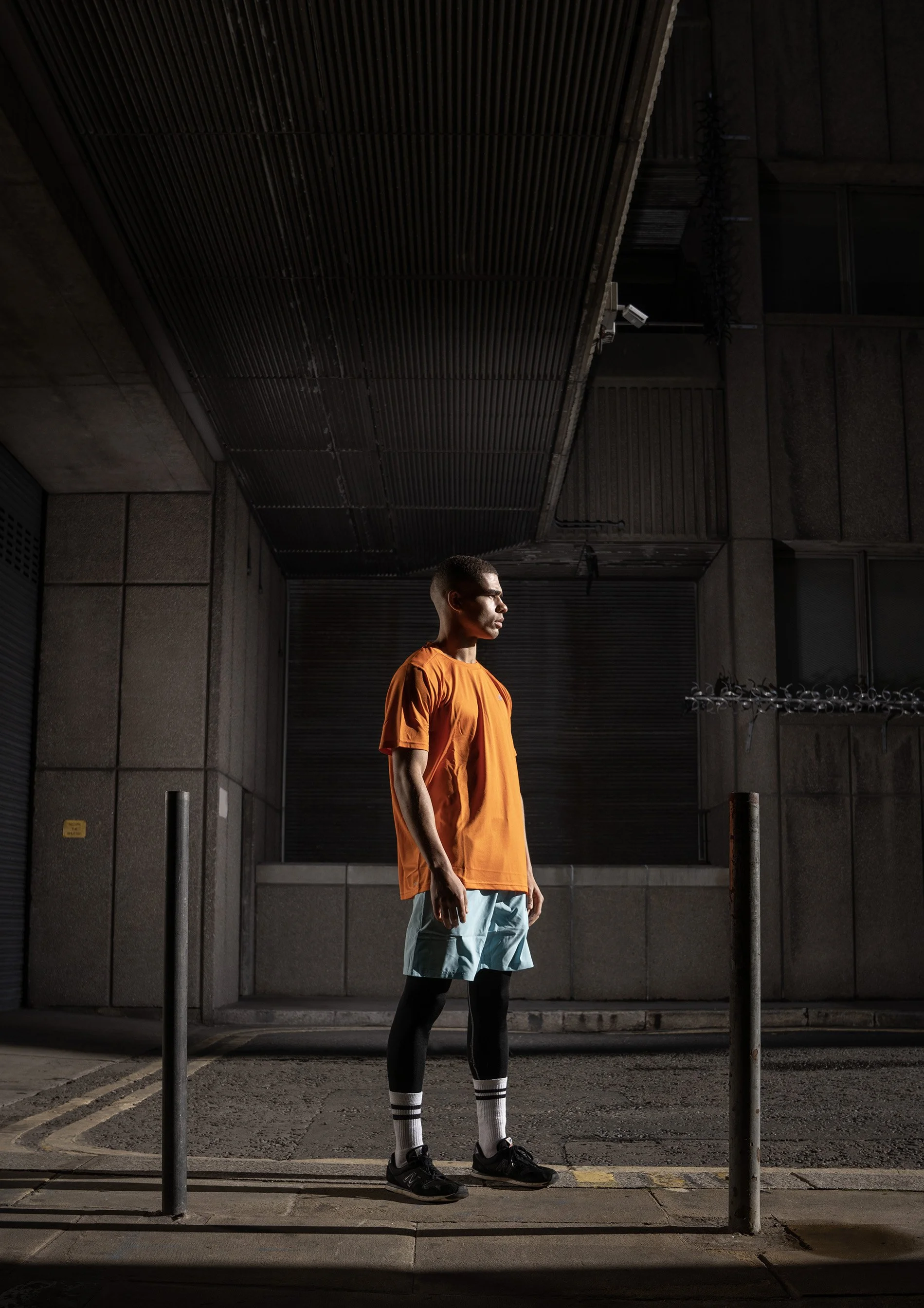A young man squats in the foreground of a brutalist urban scene, lit dramatically by the harsh sunlight. dynamic sports photography by Steve Bright