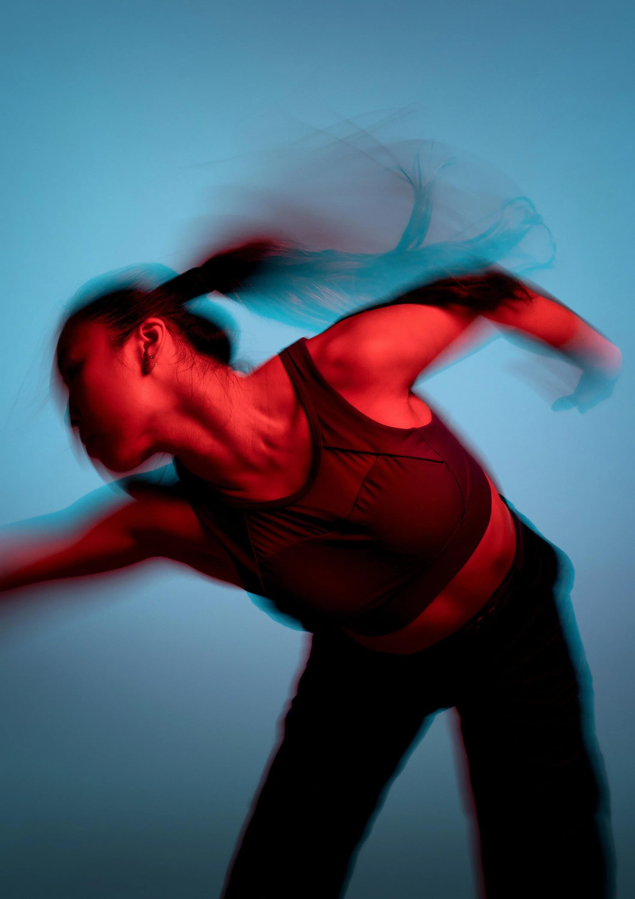 A woman in athletic wear performing a dynamic dance move, captured with motion blur against a blue background, illuminated with red lighting. dynamic sports  portrait photography by Steve Bright.
