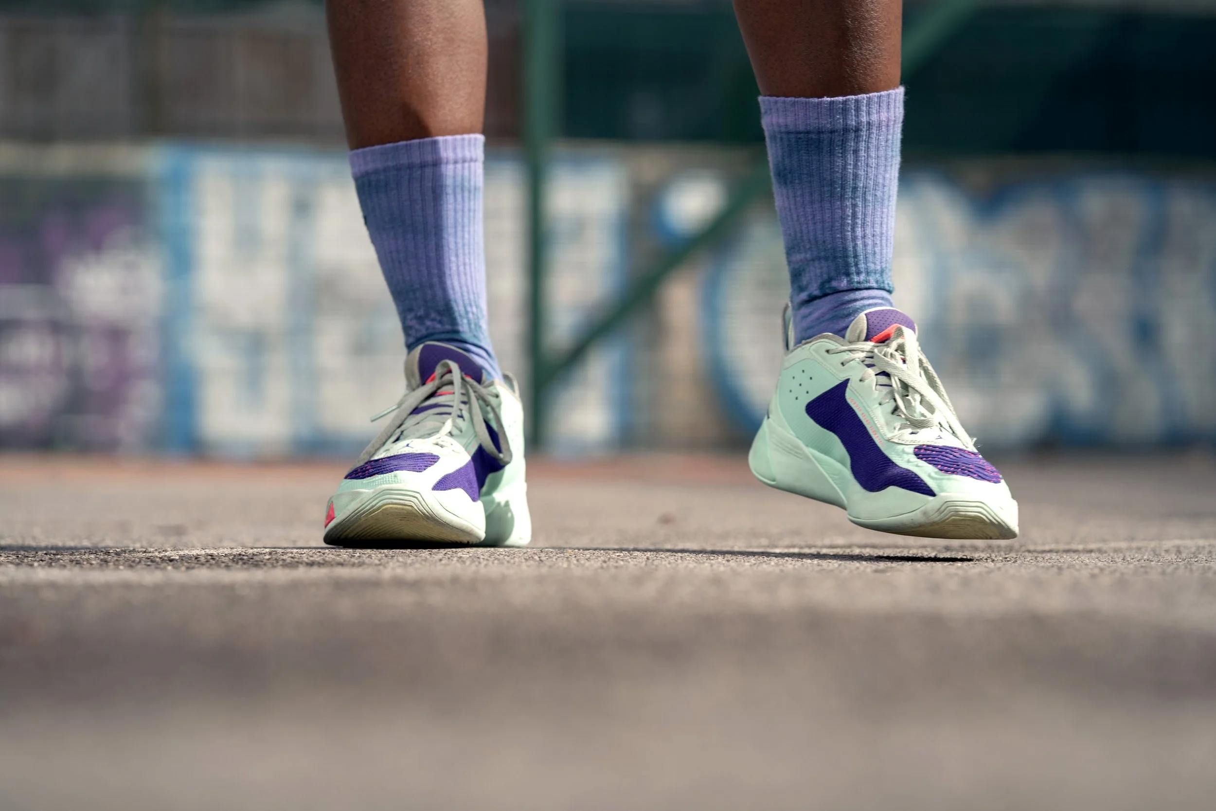 Close-up of a person's feet wearing purple and white sneakers, purple athletic socks, and standing on a rough outdoor court with a blurred graffiti wall in the background. Dynamic sports portrait photography by Steve Bright