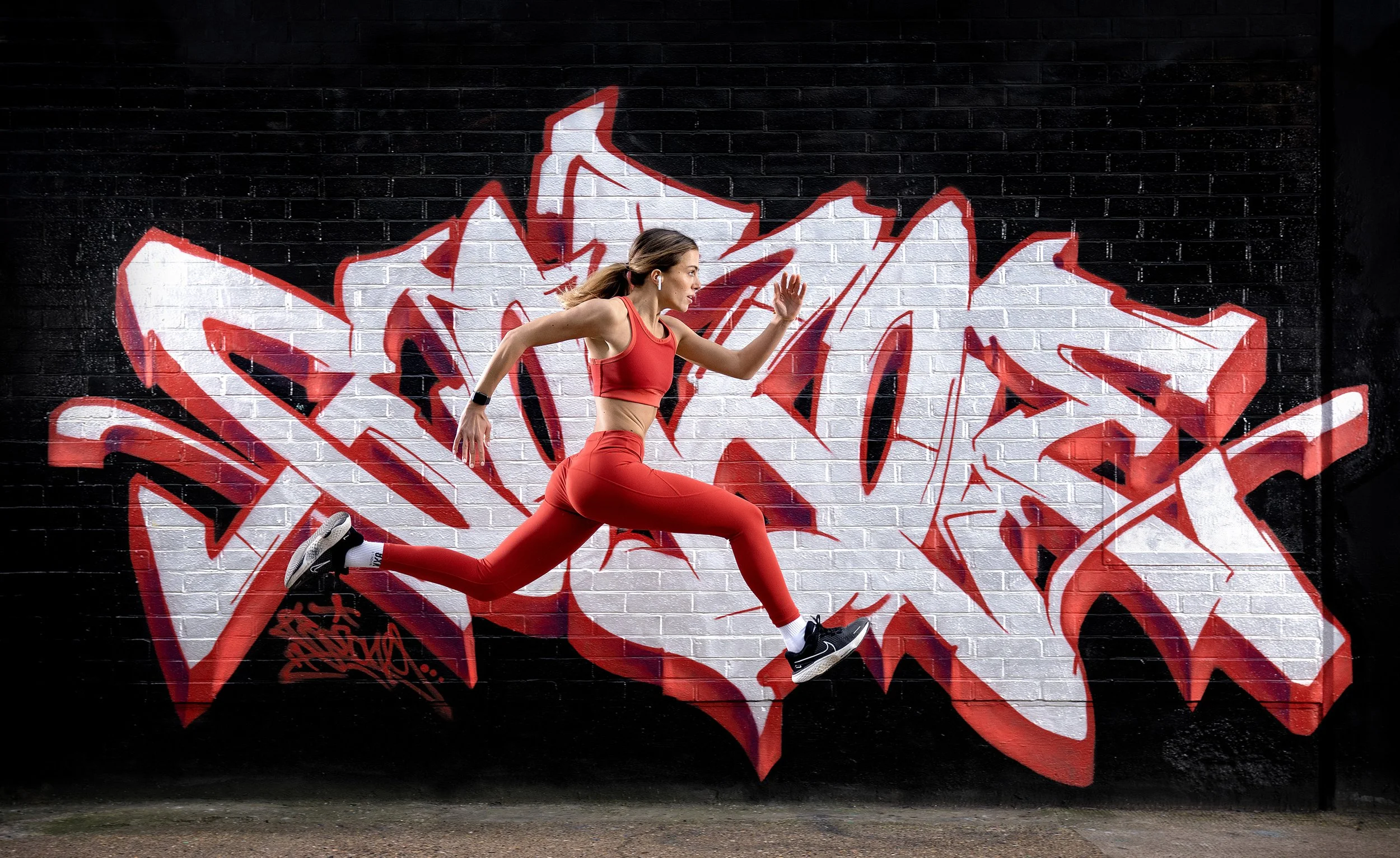 A female runner is caught mid stride as she runs at full speed in front of a graffiti mural. Her red outfit matches the red mural. dynamic sports portrait photography by Steve Bright