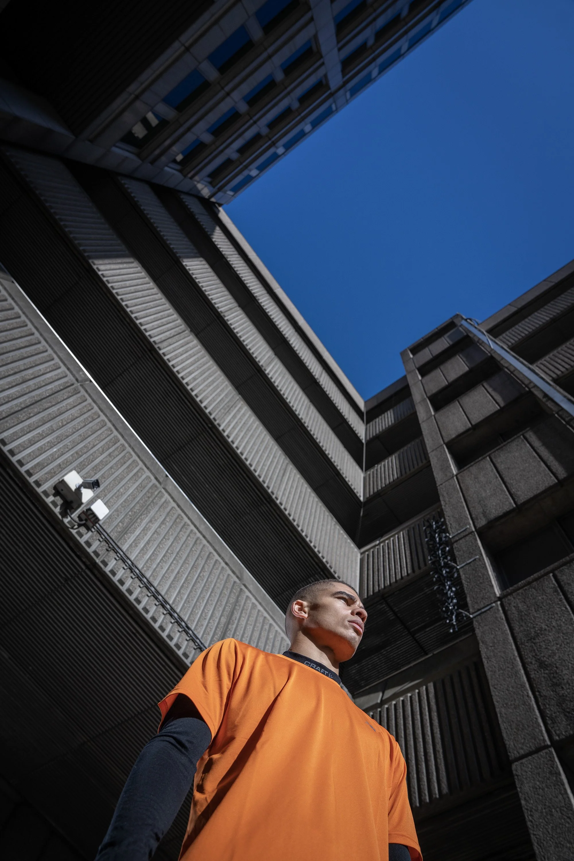 A young man in an orange t shirt is seen from below, with imposing concrete architecture towering above him. dynamic sports portrait photography by Steve Bright