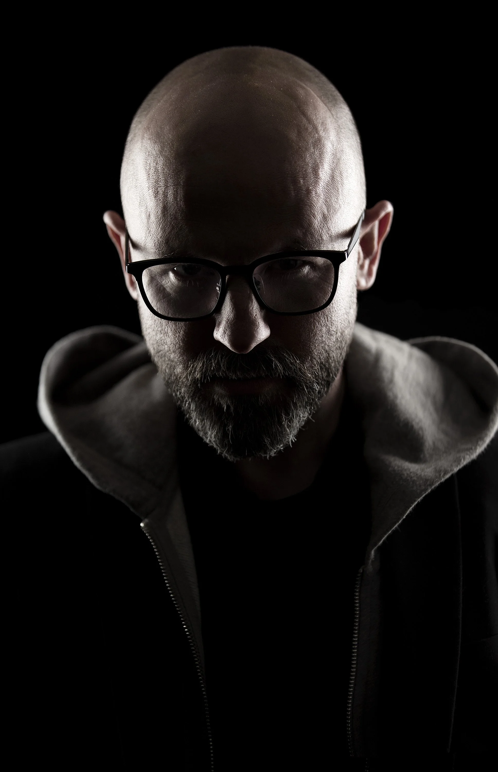 Moody shot of a bearded man in glasses, his face in darkness, wearing a hoodie. commercial portrait photography by Steve Bright