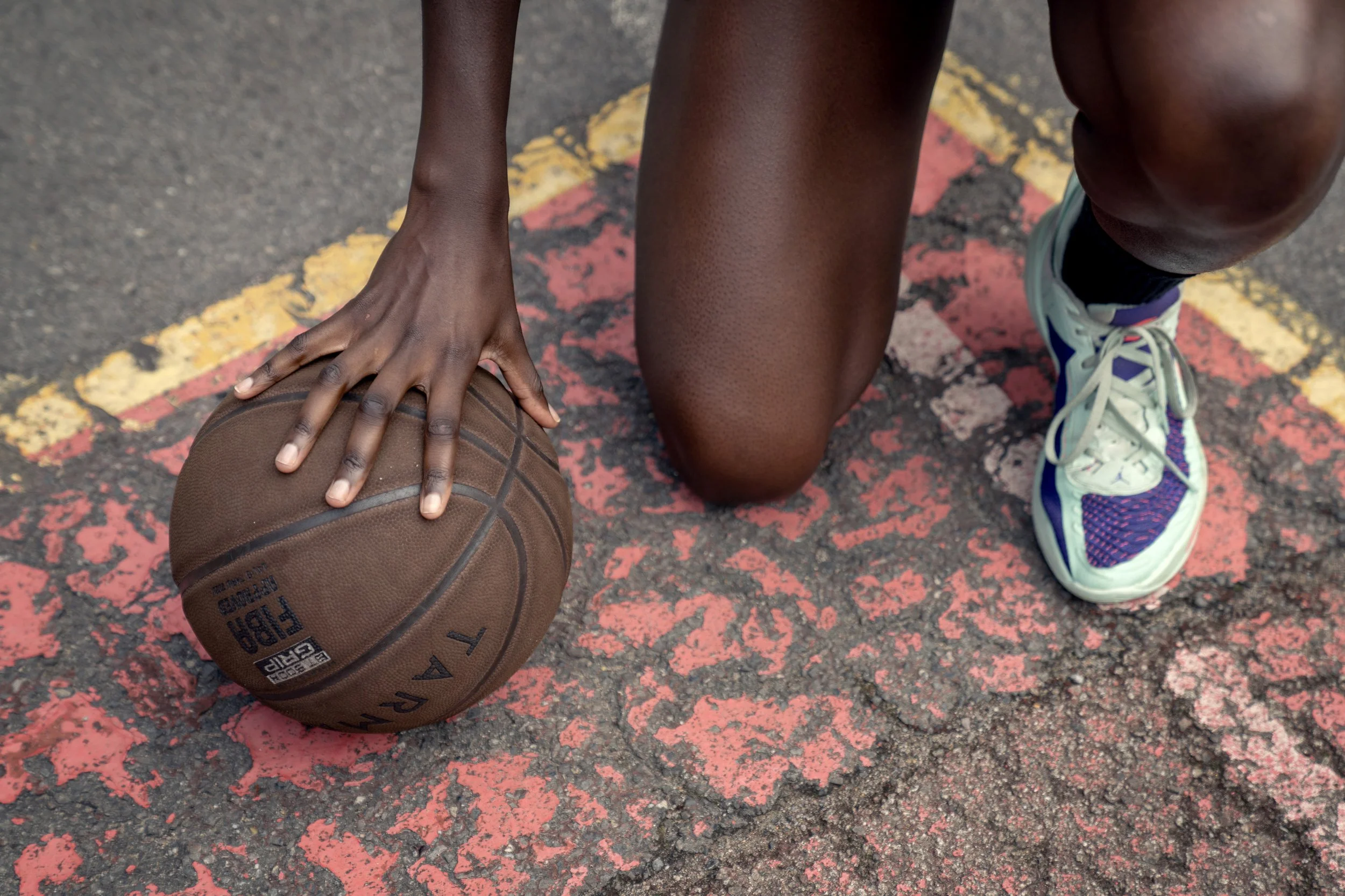 Close-up of a person with dark skin kneeling on a worn outdoor basketball court, preparing to bounce a brown basketball with black markings, wearing colorful sneakers. Dynamic sports portrait photography by Steve Bright