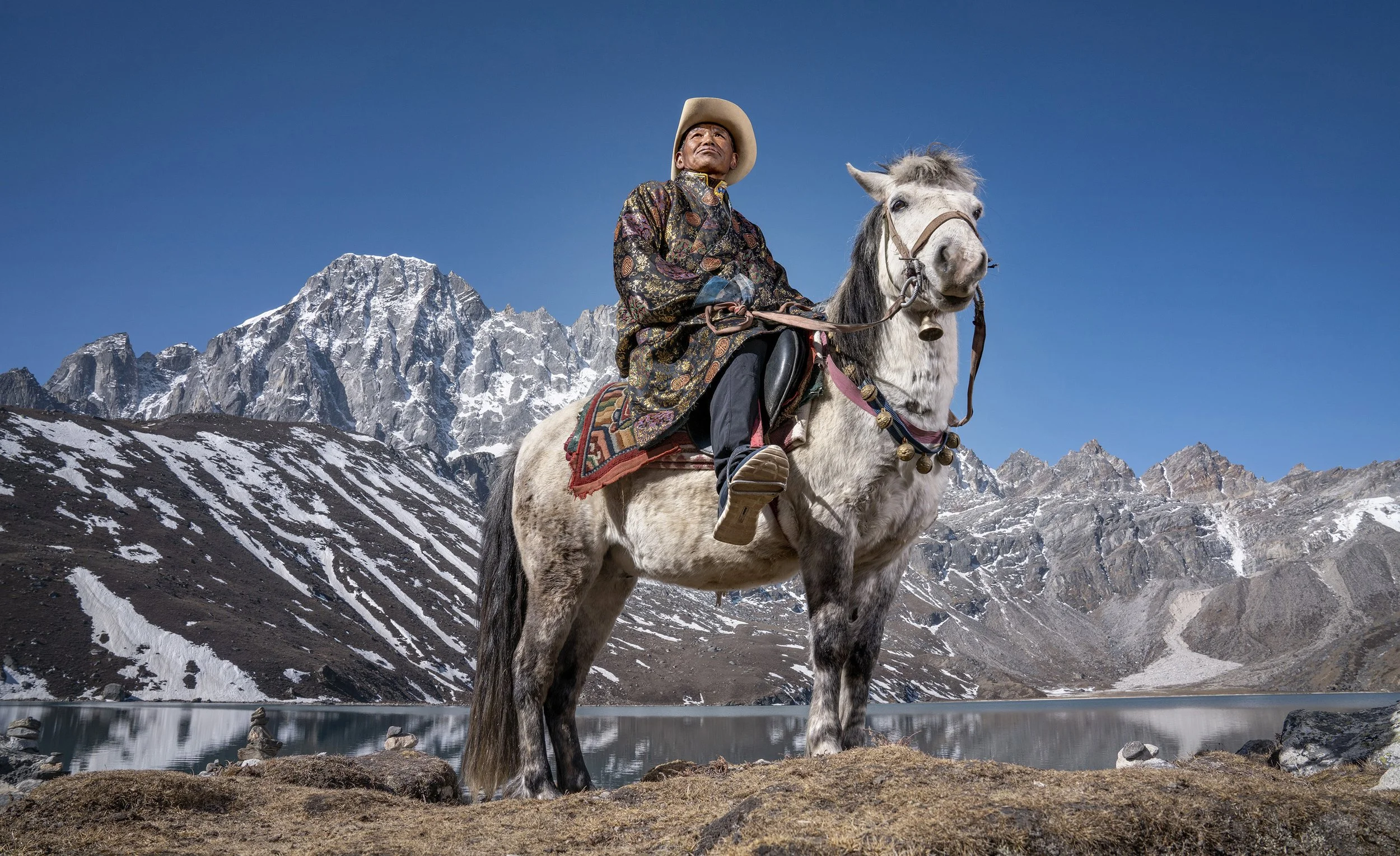 An elderly sherpa man in a long patterened jacket and a cowboy hat sits on a horse in front of Gokyo Lake in Nepal with mountains in the background. commercial portrait photography by Steve Bright.