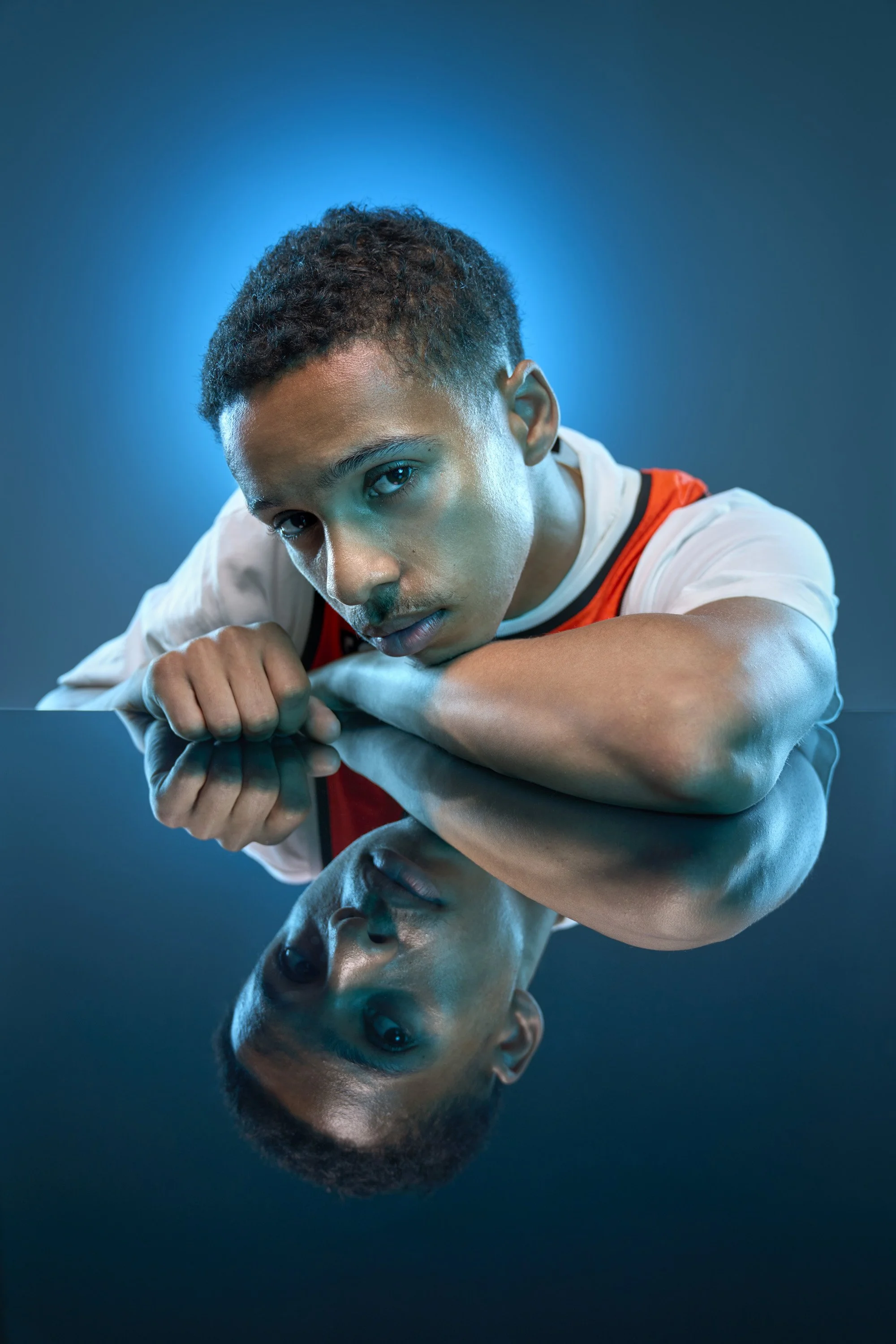 A young man wearing an ornage vest over a white t shirt rests his arm on a reflective surface, we see his reflection as well as his actual head and shoulders.t
