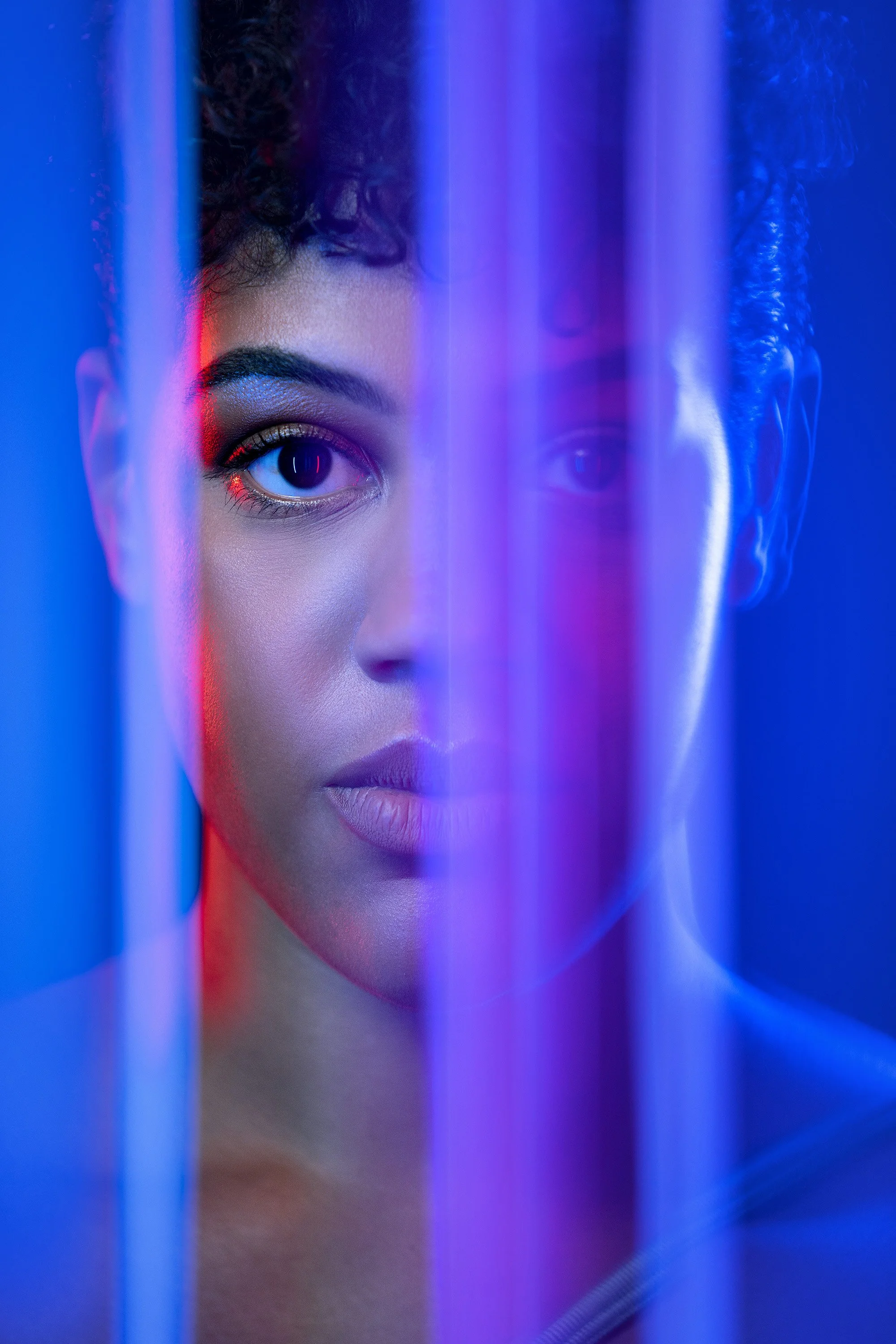 Graphic and stylised portrait of a young woman staring through vertical neon lit bands of glass, her face distorted through the different vertical planes. stylised commercial portrait photography by Steve Bright