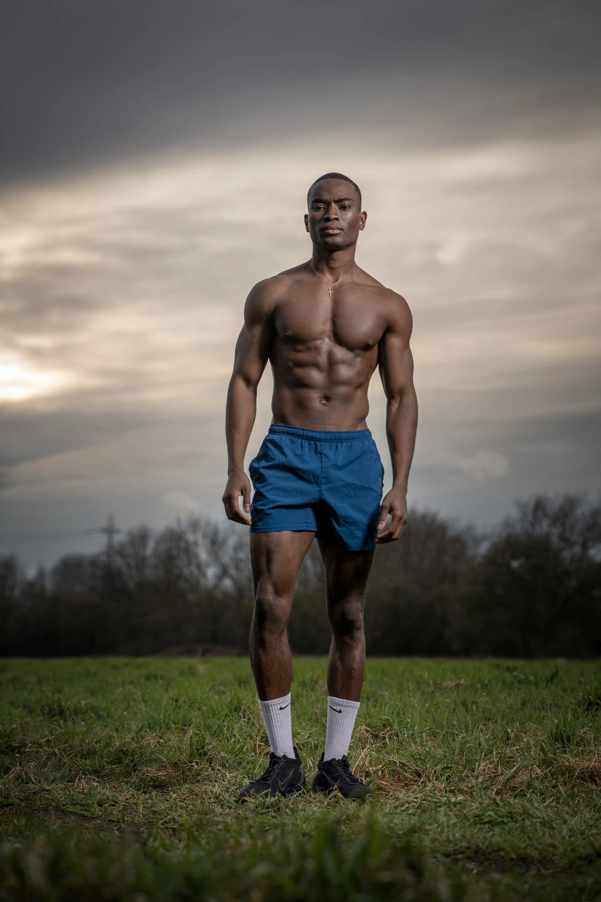 A shirtless young man with a muscular build standing outdoors on grass, wearing blue shorts, white Nike socks, and black sneakers, with an overcast sky in the background. dynamic sports portrait photography by Steve Bright