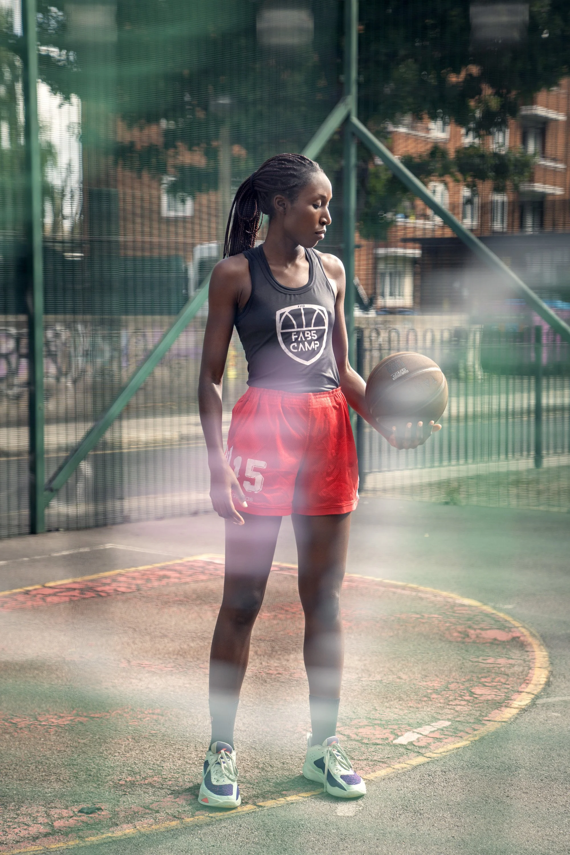 A young woman standing on an outdoor basketball court, holding a basketball, viewed through a chain-link fence which has been sprayed with graffiti. dynamic sports  portrait photography by Steve Bright