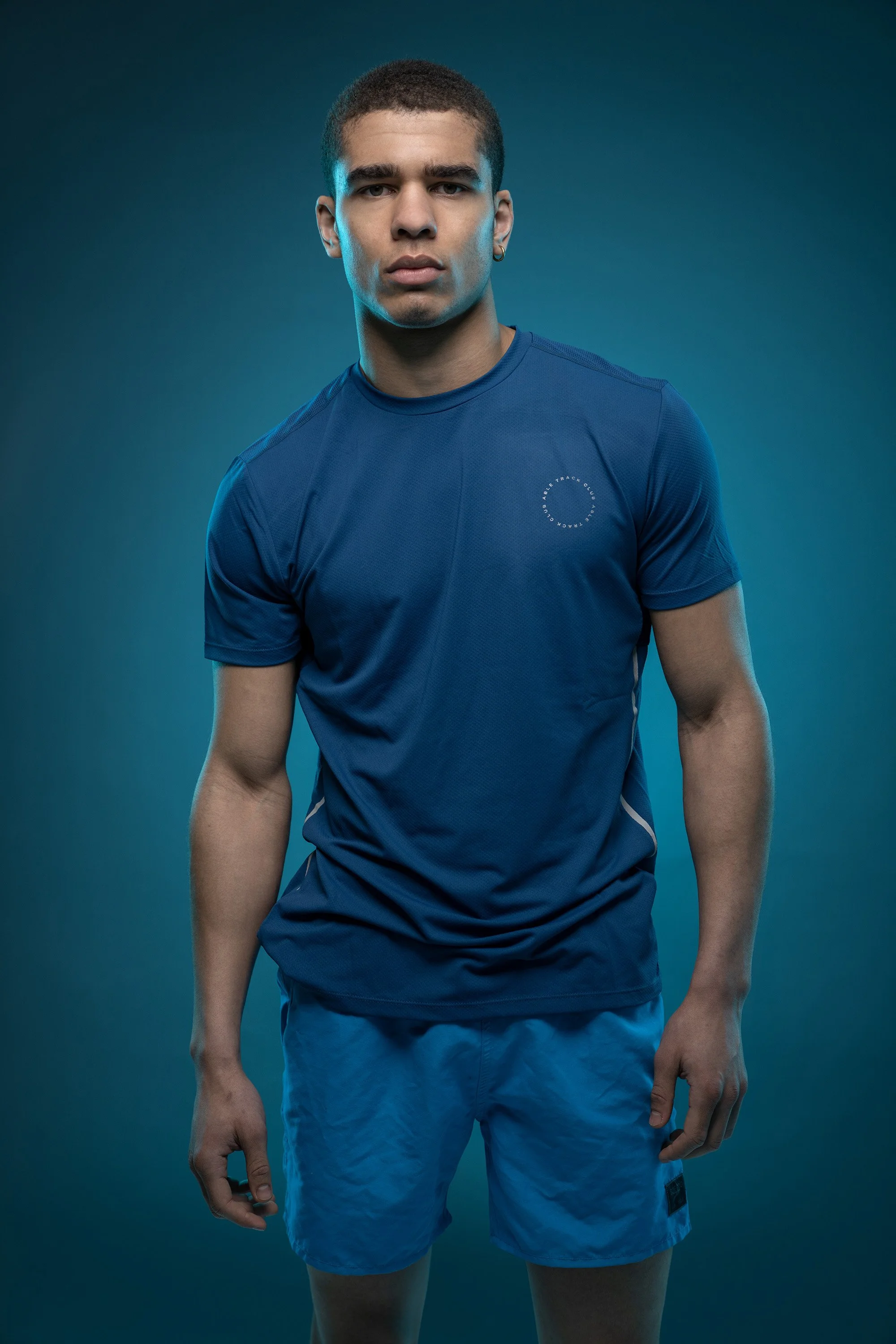 A handsome young boxer stares into the camera, his face sidelit with blue-green neon lighting. He wears a blue t shirt and blue shorts. commercial portrait photography by Steve Bright