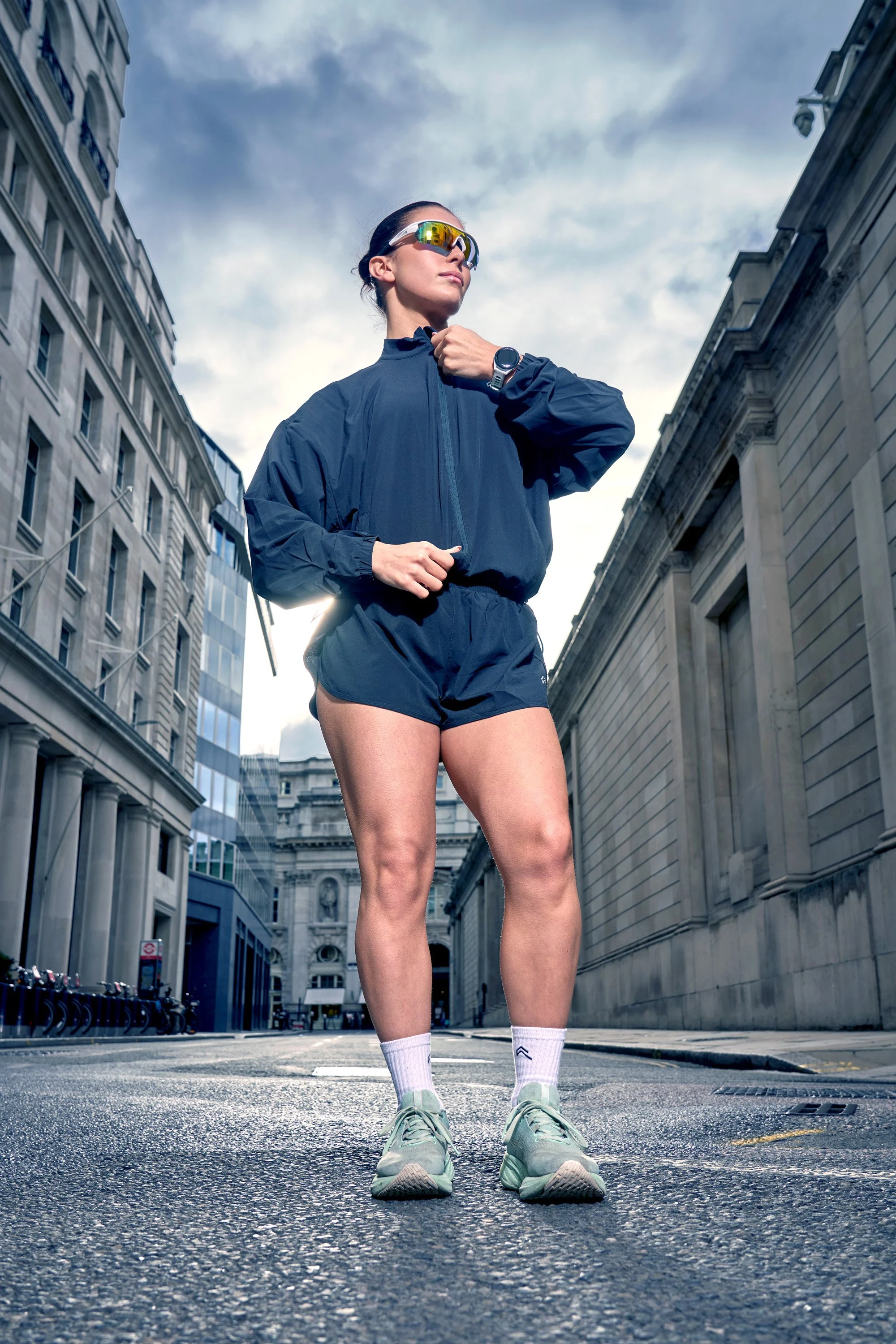 A young woman in fitness gear stands on a city street doing up the zip of her running jacket. dynamic sports  portrait photography by Steve Bright