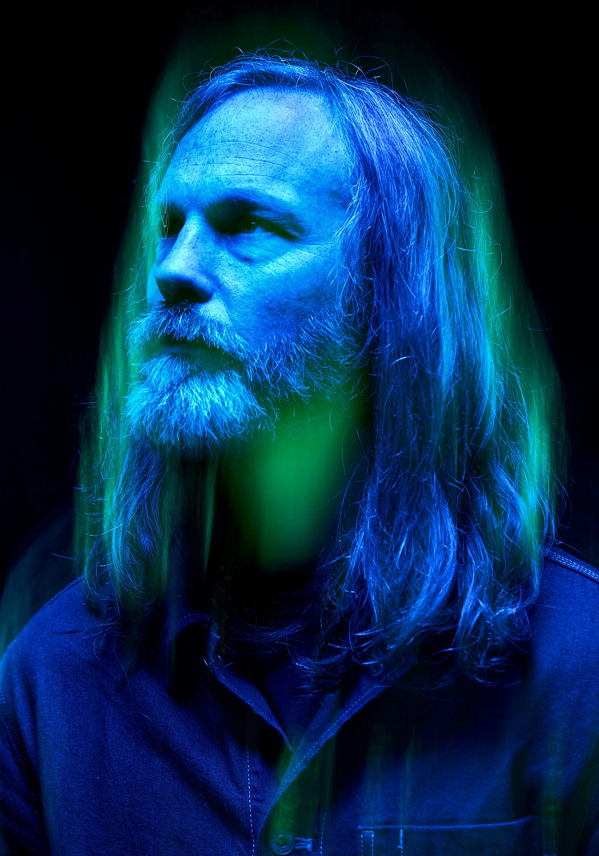 A man with long hair and a beard looks off camera. He is litith blue light and surrounded by a shimmering green aura