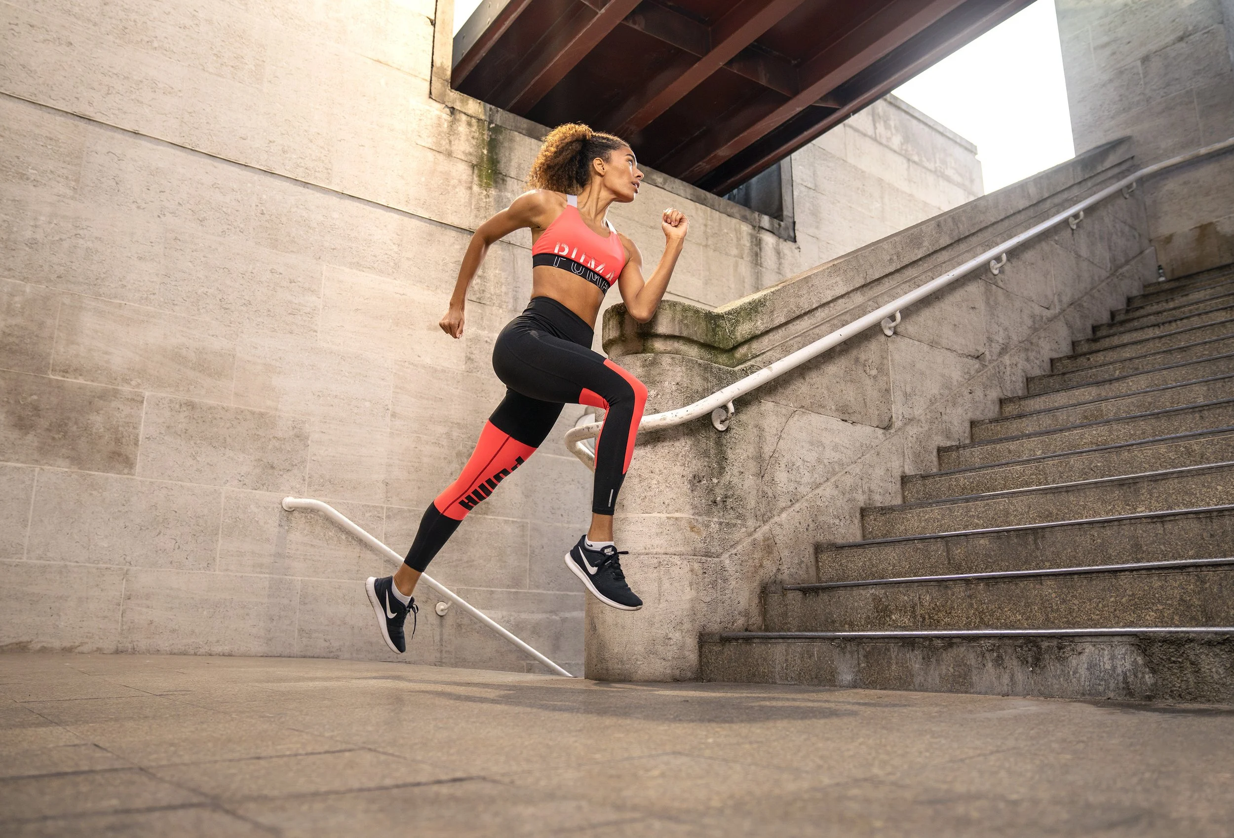 A woman in athletic clothing is running up a concrete staircase outdoors, with a wall and part of a bridge overhead. dynamic sports portrait photography by Steve Bright