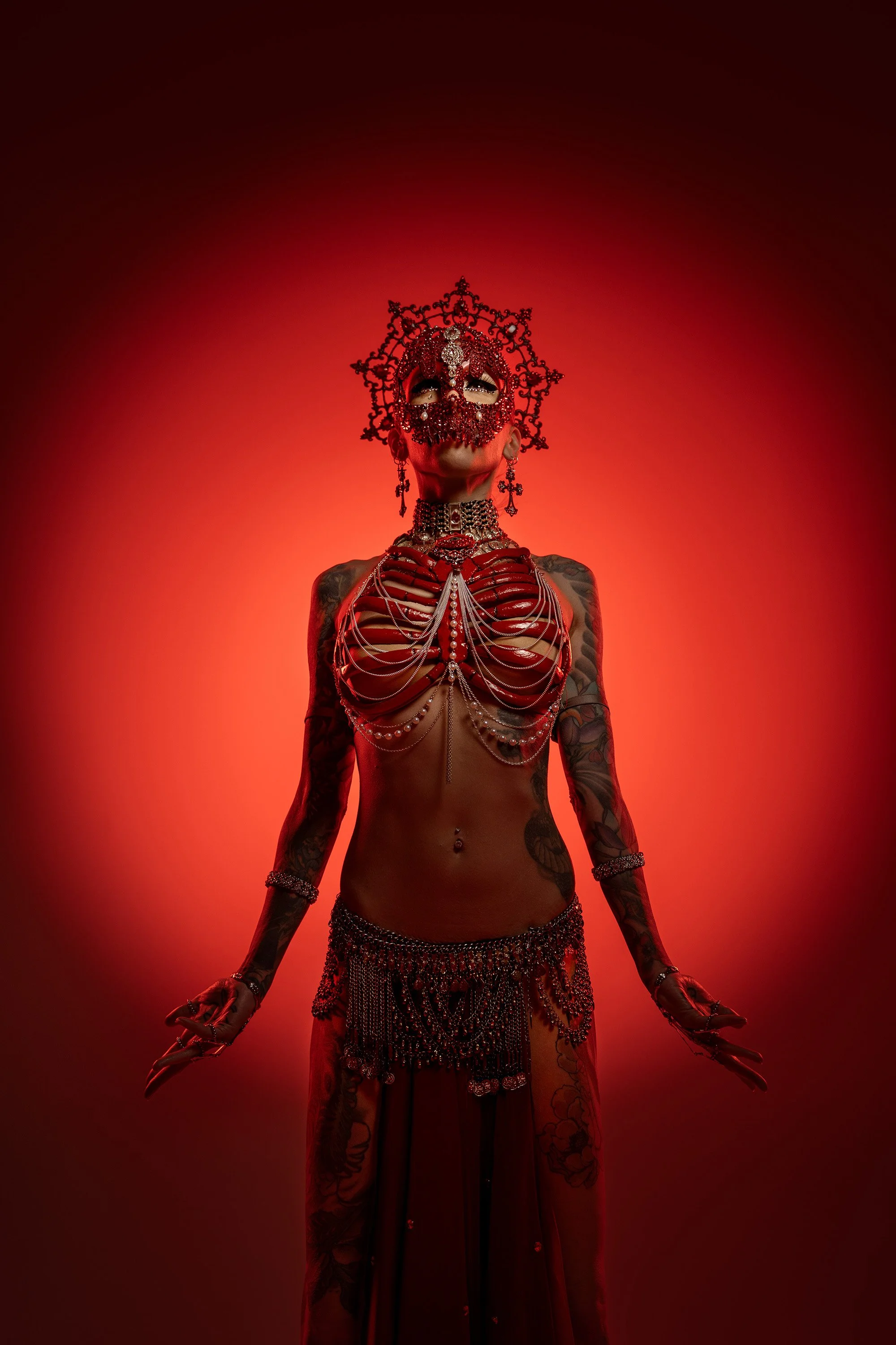 The performer and dancer Leah Debrincat stands, moodily lit, in front of an illuminated red circle background. She wears a jewelled skull headpiece and a chestpiece that looks like a ribcage. entertainment portrait photography by Steve Bright, London