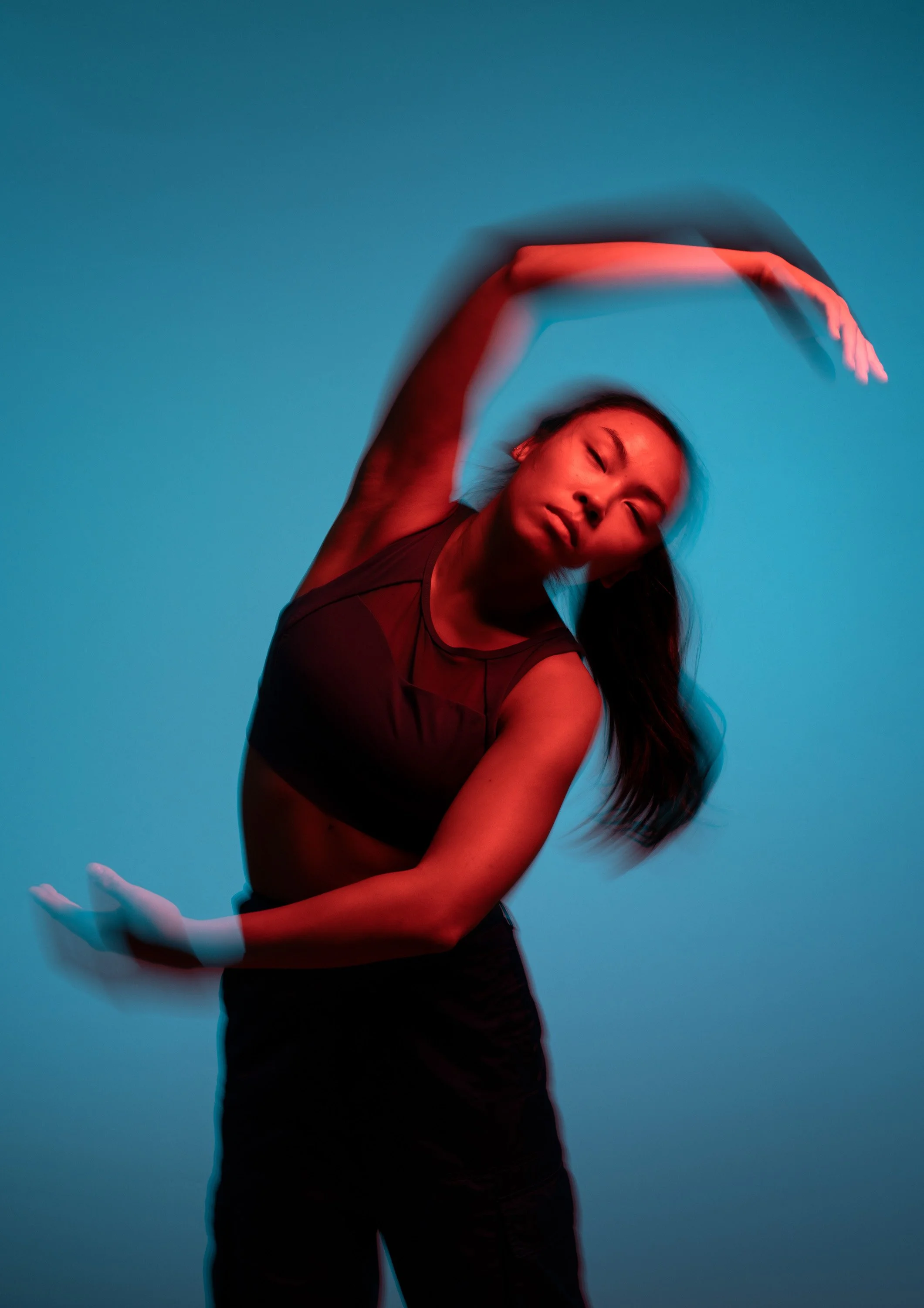 A woman in athletic wear performing a dynamic dance move, captured with motion blur against a blue background, illuminated with red lighting. dynamic sports  portrait photography by Steve Bright.