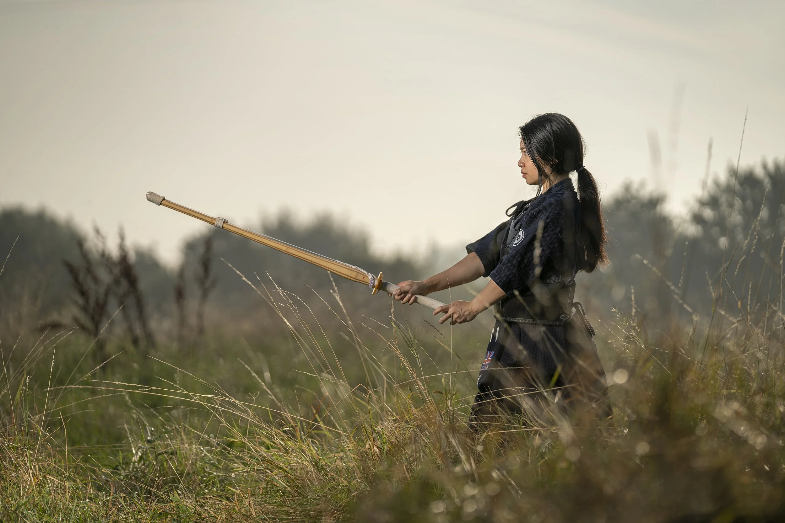 A woman stands in a grassy field holding a wooden sword, dressed in traditional kendo armor, , with a focused expression. dynamic sports portrait photography by Steve Bright