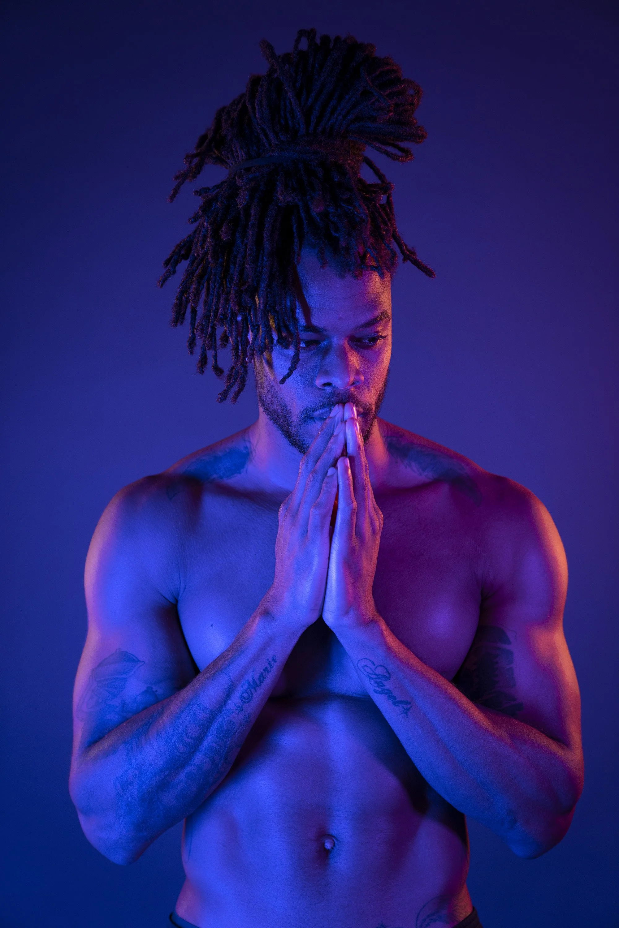 A man with dreadlocks stares thoughtfully over his clasped hands, lit with blue and pink neon light. stylised commercial portrait photography by Steve Bright