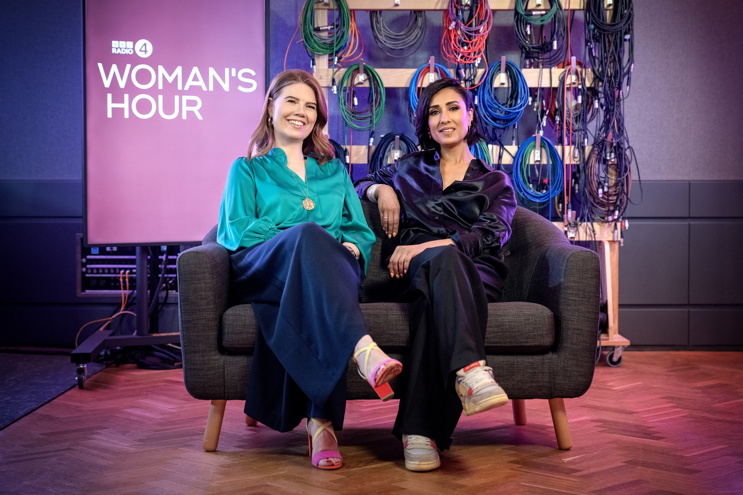 Woman's Hour Presenters Anita Rani and Nuala McGovern sitting on a small sofa in a BBC recording studio, in front of a rack of cables and a screen with the words Woman's Hour on it. dynamic entertainment portrait photography by Steve Bright London