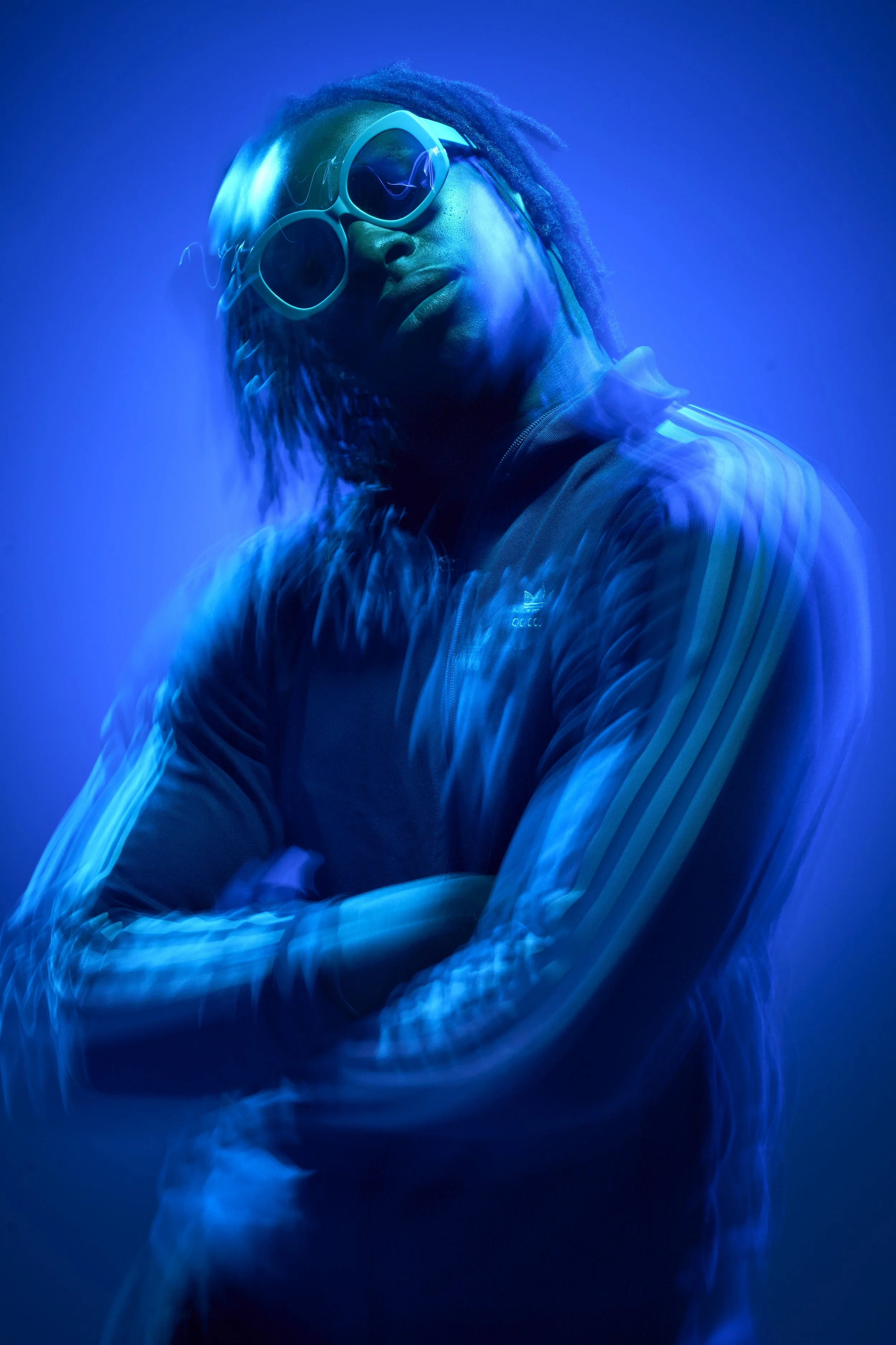 A young Black man with short dreadlocks, wearing a blue Adidas tracksuit top and matching sunglasses, cocks his head and looks into camera. The lighting is blue, blurry and dreamlike, only his face is crisp and sharp.