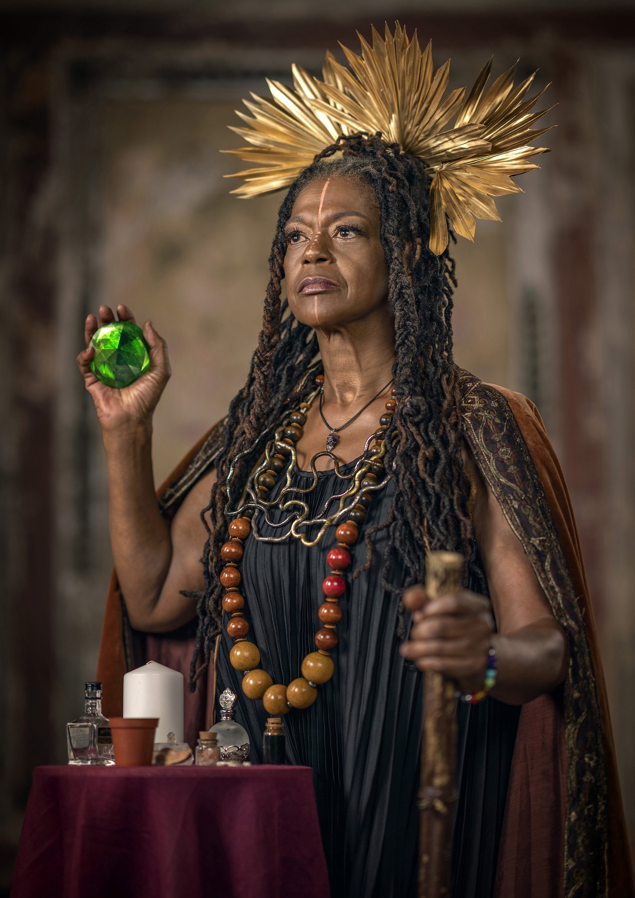 An older Black woman holding a large green jewel and standing in front of a small altar poses in a modern reimagining of a regal outfit, in a modern Afrofuturist version of a royal portrait. commercial portrait photography by Steve Bright