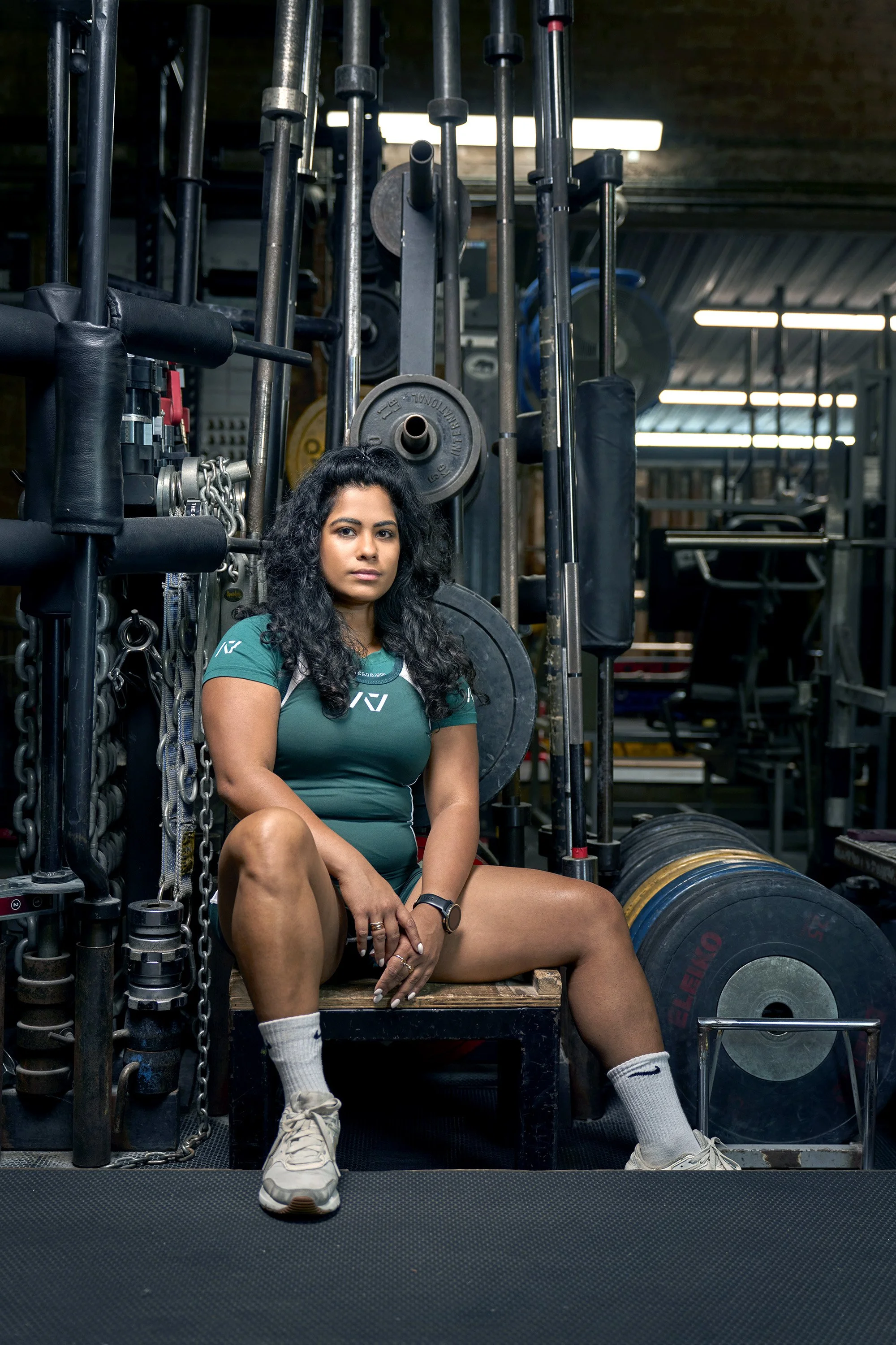 A female powerlifter sitting in a gym, surrounded by weightlifting equipment. dynamic sports portrait photography by Steve Bright