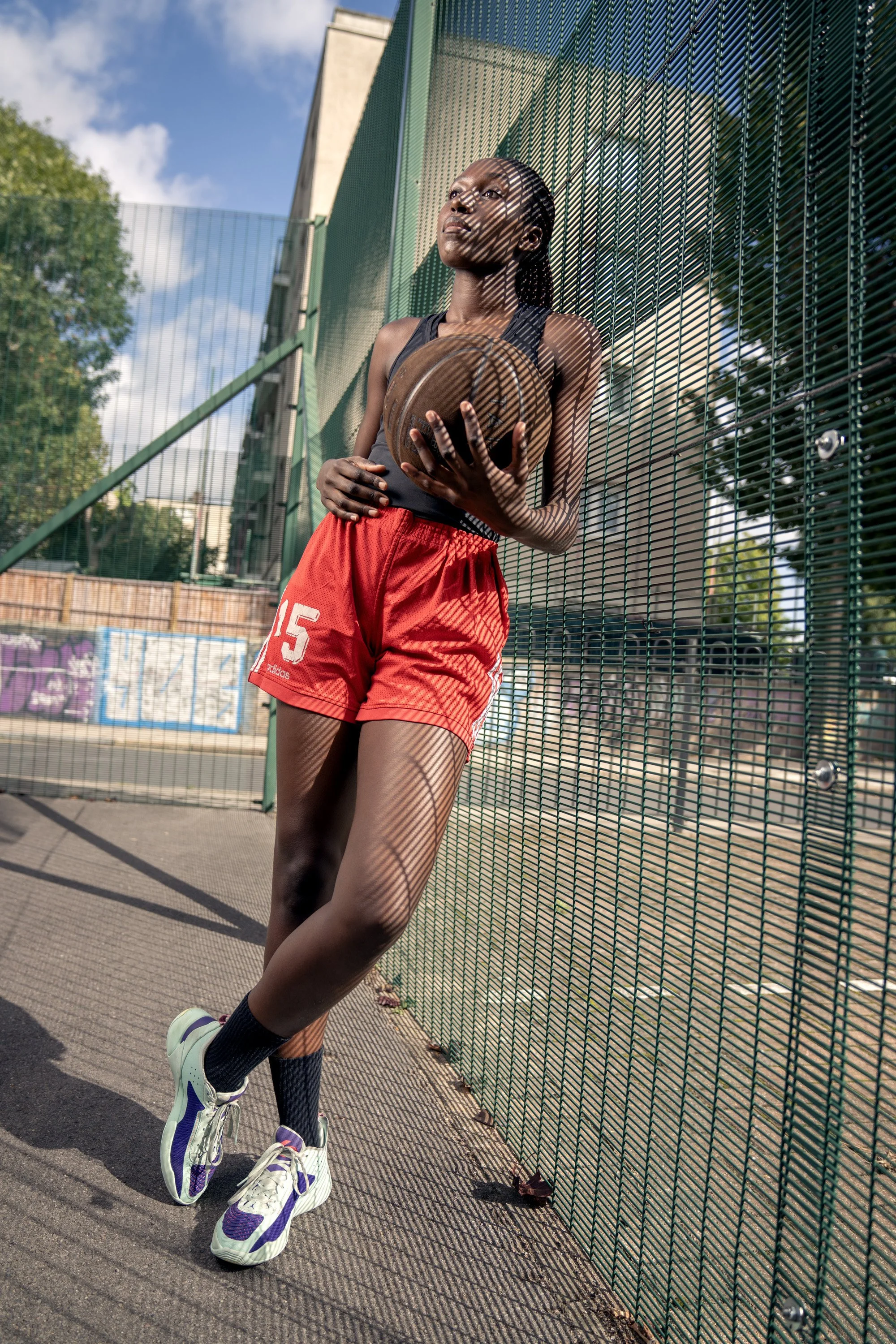 A young woman standing on an outdoor basketball court, leaning against a wire fence with the shadows of the fence cast on her face and body, holding a basketball, dynamic sports  portrait photography by Steve Bright