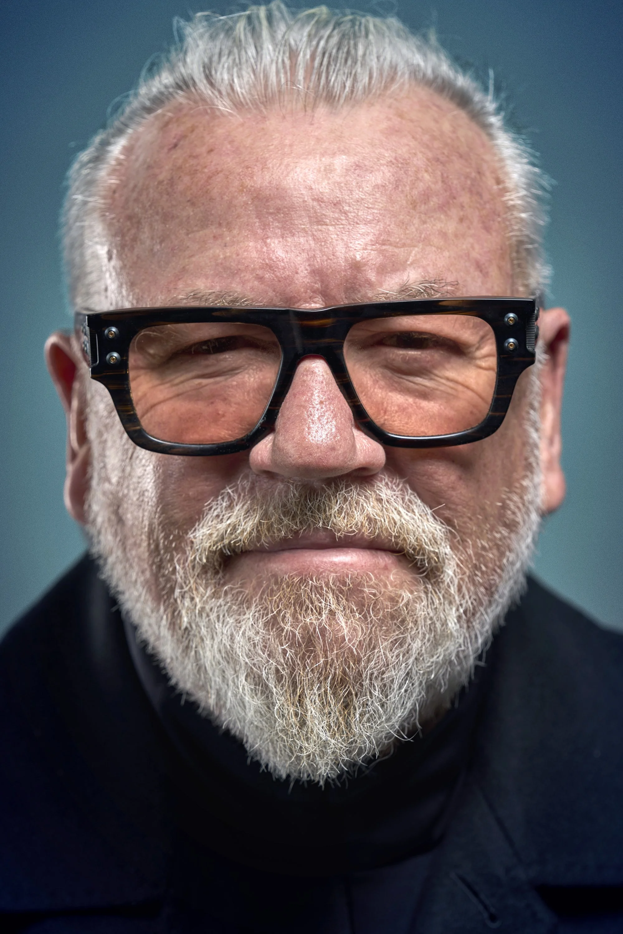 The actor Ray Winstone smiles into the camera wearing orange tinted thick framed glasses, with a blue background behind him. commercial portrait photography by Steve Bright