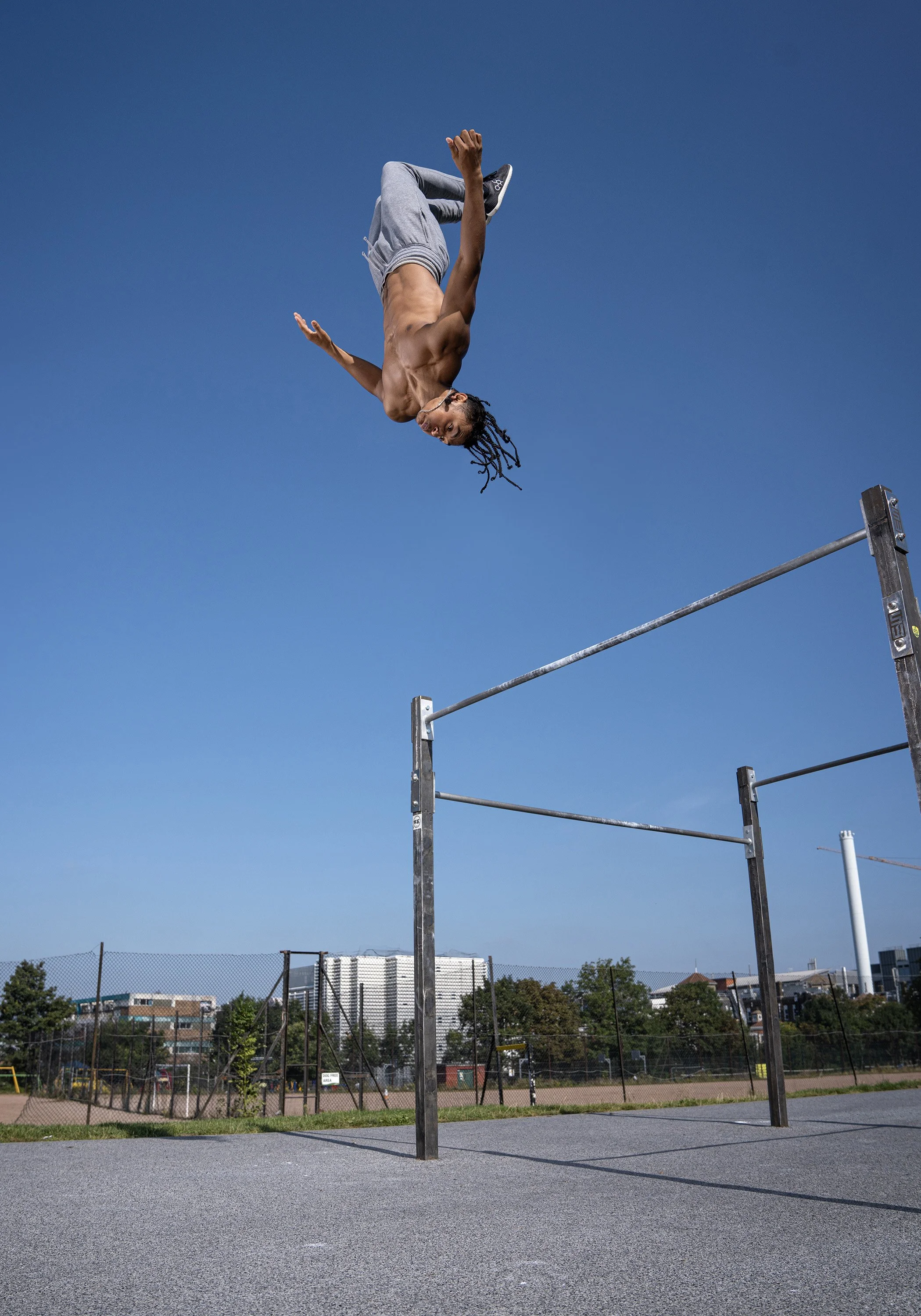 A man with dreadlocks is mid-air, backflipping off bars at an outdoor park under a clear blue sky. dynamic sports portrait photography by Steve Bright