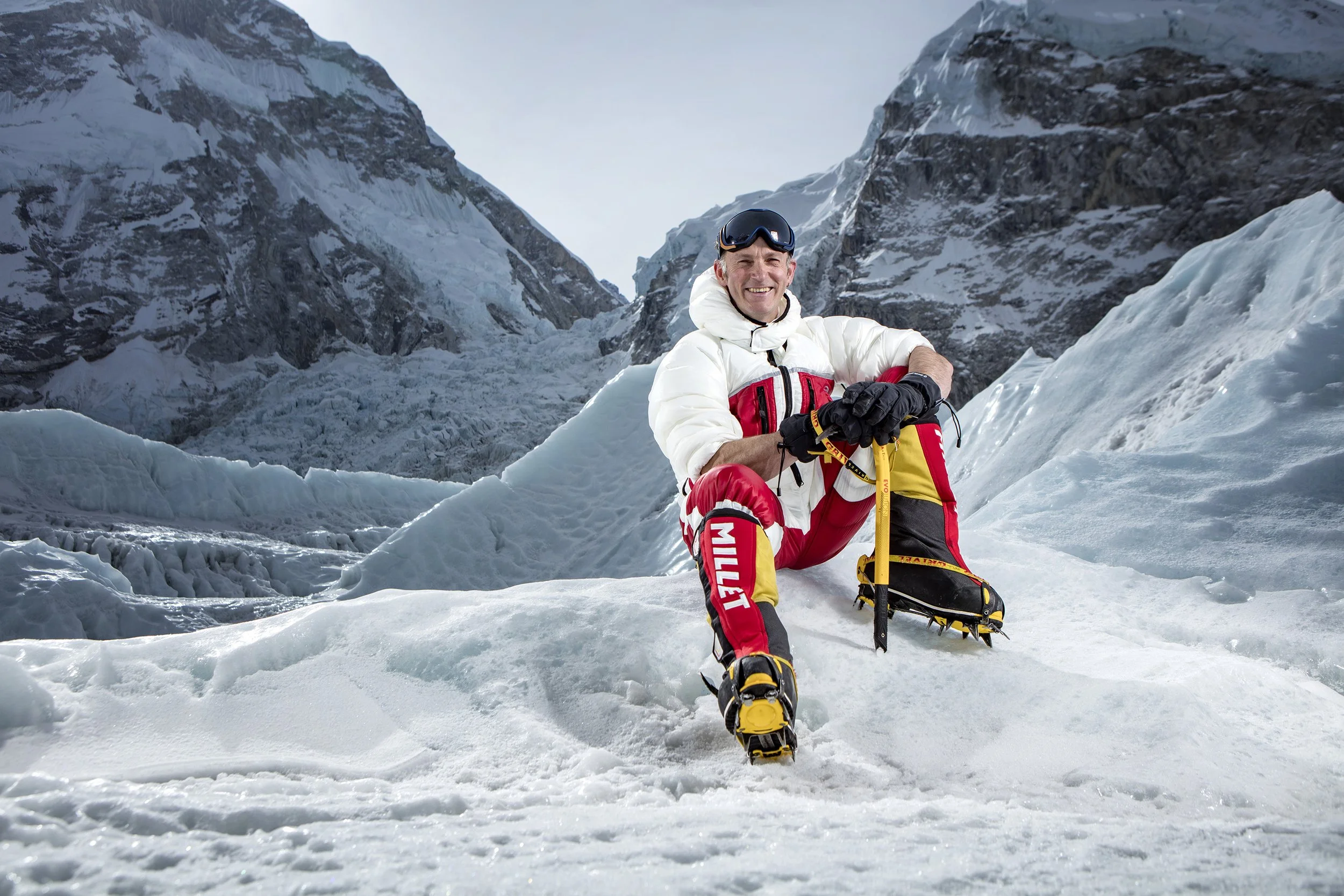 A british mountain climber sits on the lower reaches of the Khumbu Icefall on Everest, smiling at the camera. He wears a down suit, crampons and holds an ice axe. commercial portrait photography by Steve Bright