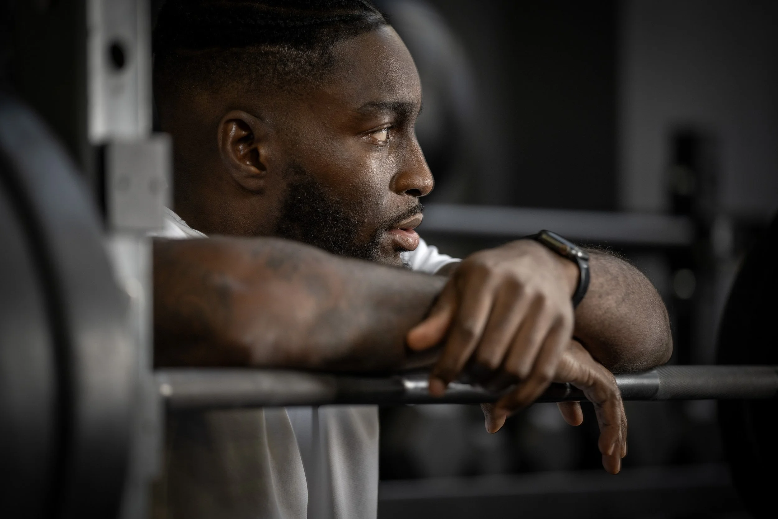 A man resting his arms on a barbell, looking thoughtfully to the side. dynamic sports portrait photography by Steve Bright
