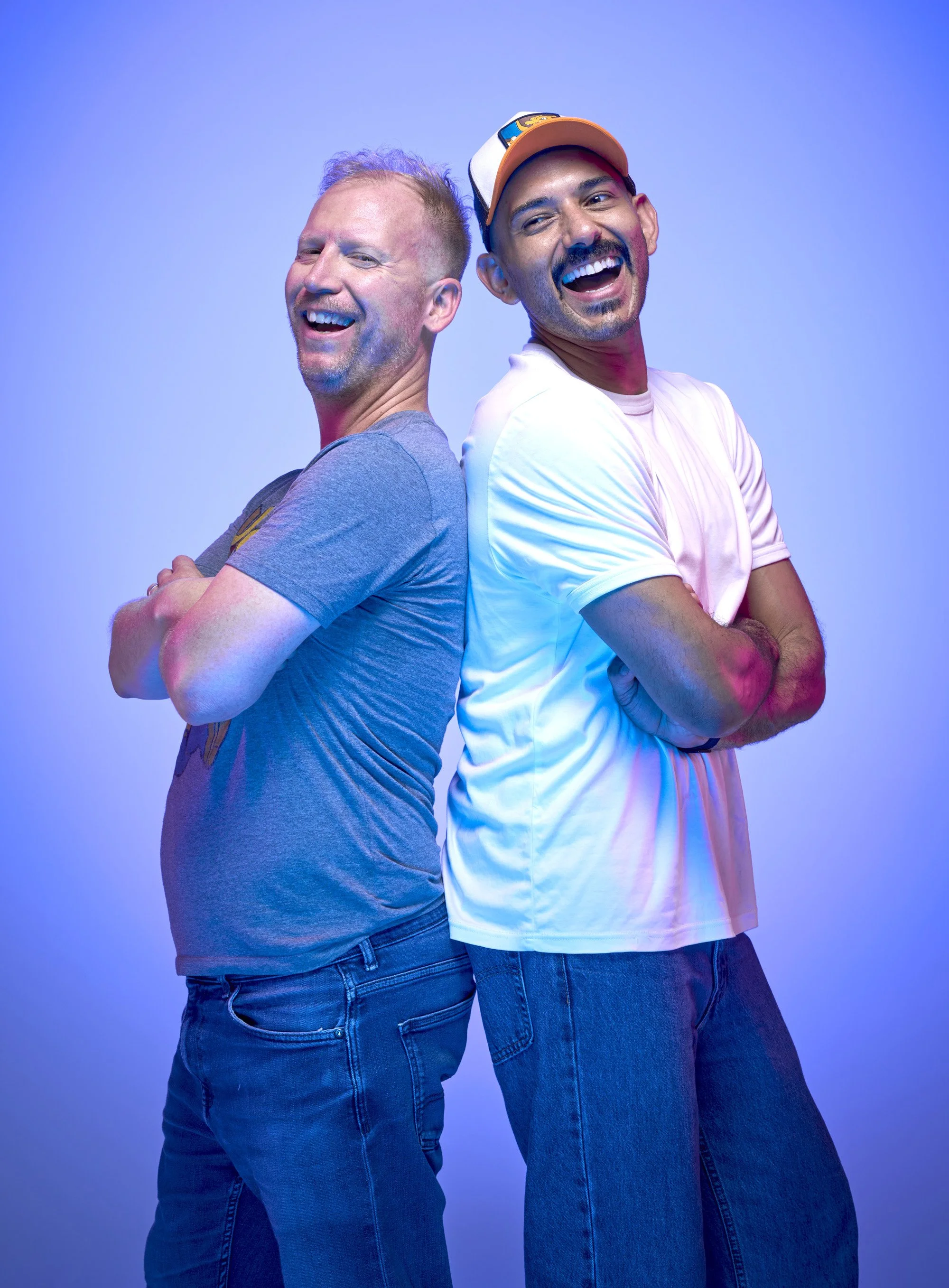 Portrait of the two hosts of the gaming podcast Out Of Sync. The two male hosts stand back to back with arms crossed, smiling at the camera. On a blue background and lit with blue and pink highlights.