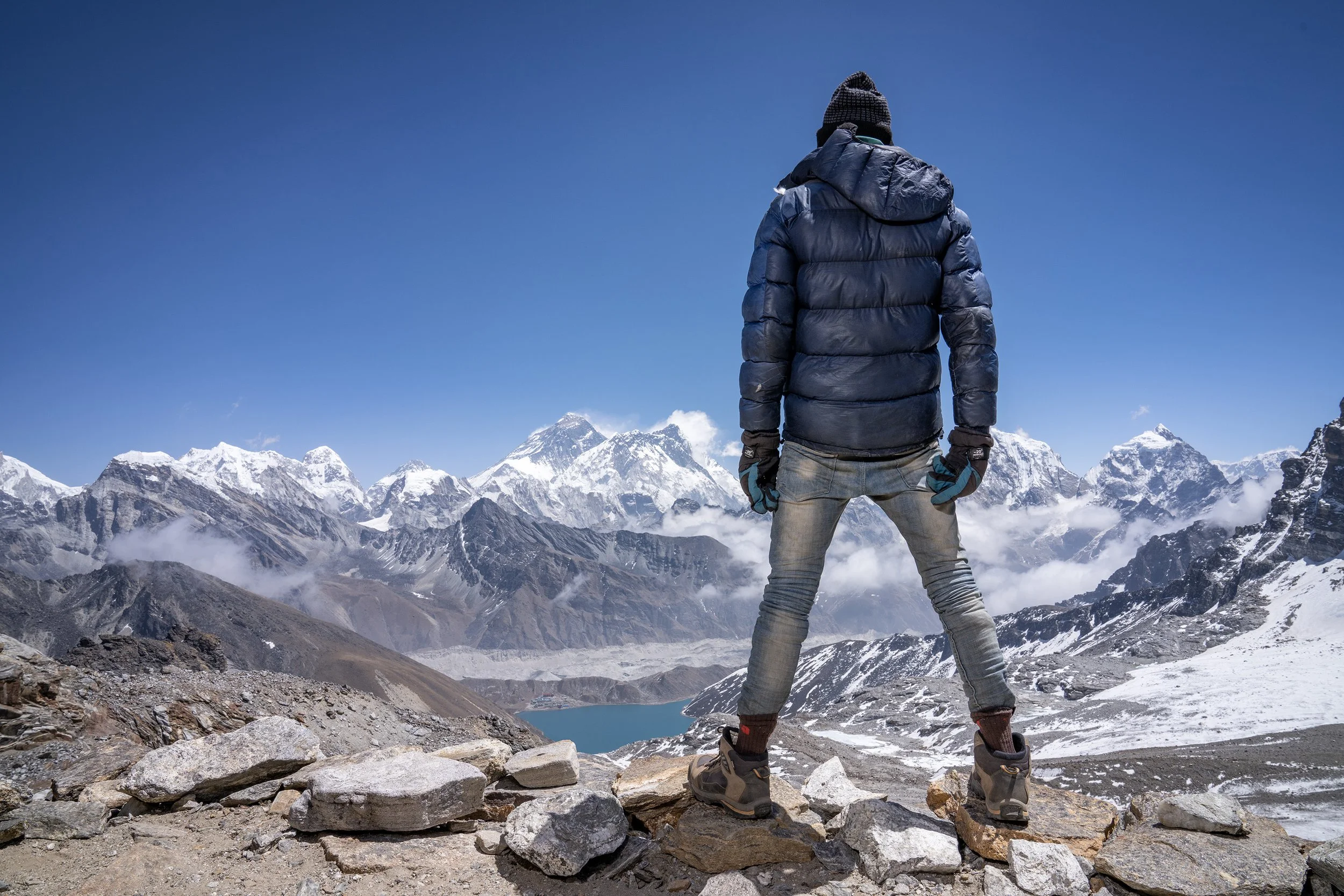 A young Nepalese porter stands at the top of a high pass in the Himalayas surveying a mountain range, including Mount Everest. He wears skinny jeans, a down jacket and hat and gloves. commercial portrait photography by Steve Bright