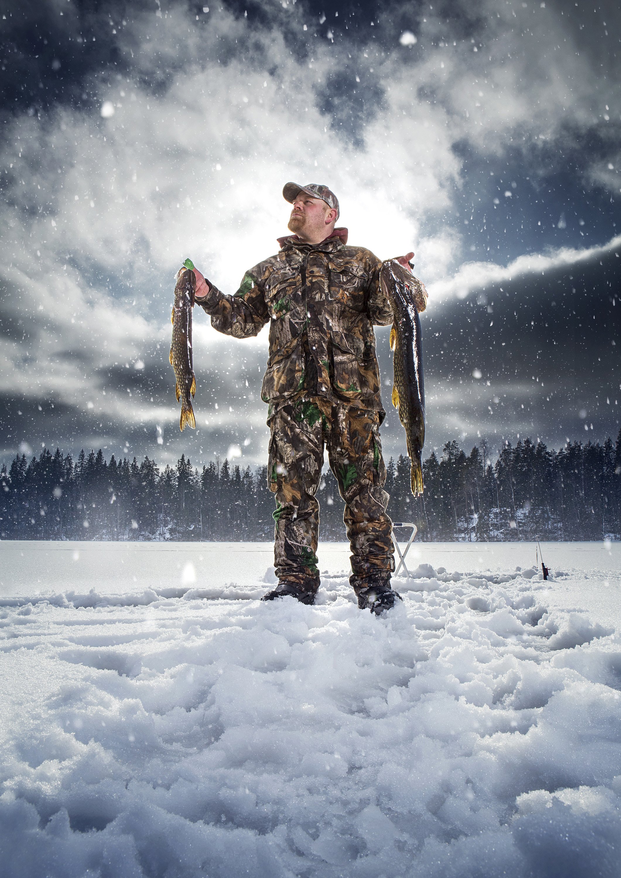An ice fisherman stands on a frozen lake in Finlanddressed in camo cold weather gear, holding two large fish he has caught. commercial portrait photography by Steve Bright
