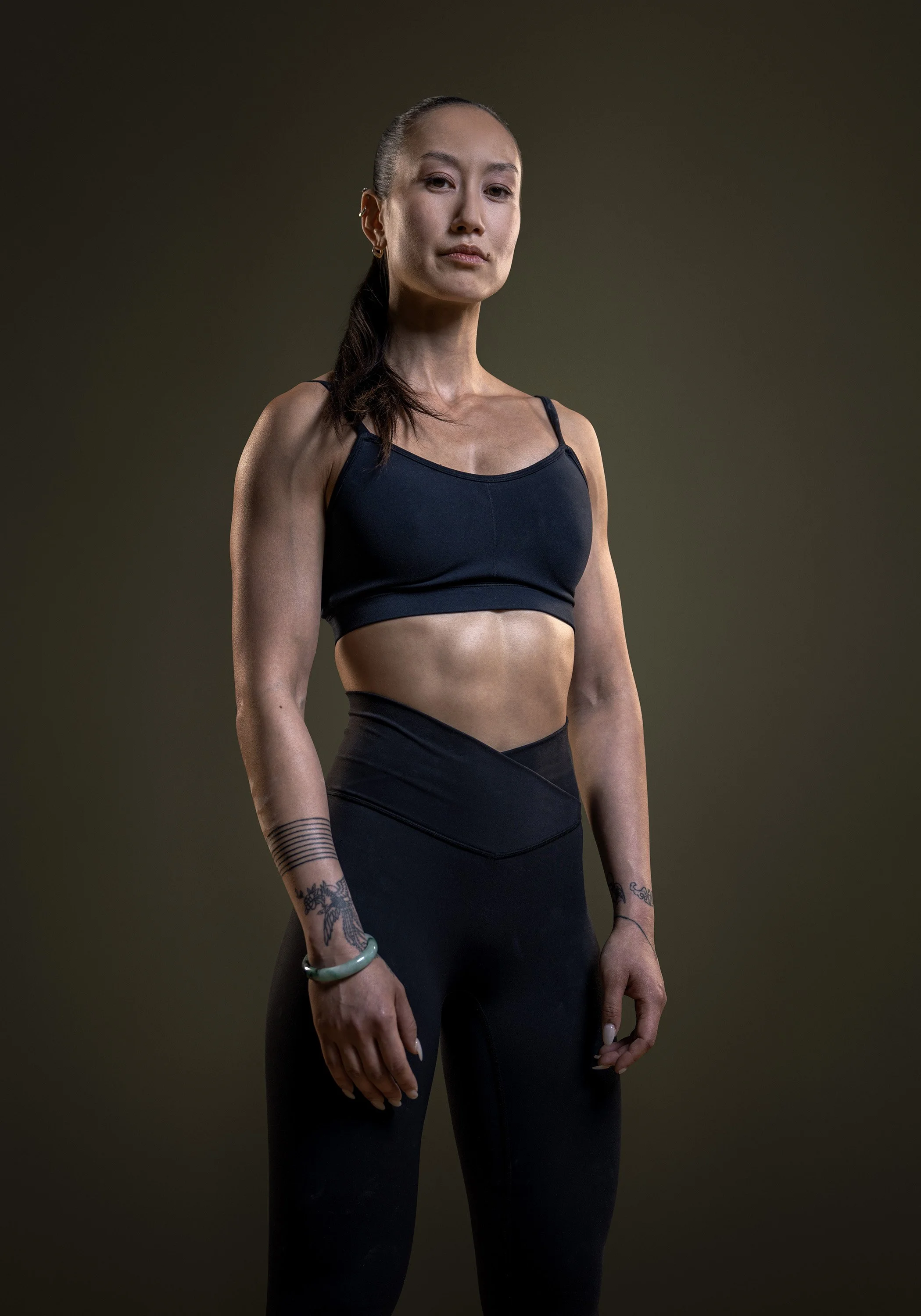A young, very fit asian woman stands, wearing black fitness clothing, looking confidently into camera, on a dark green background. commercial portrait photography by Steve Bright