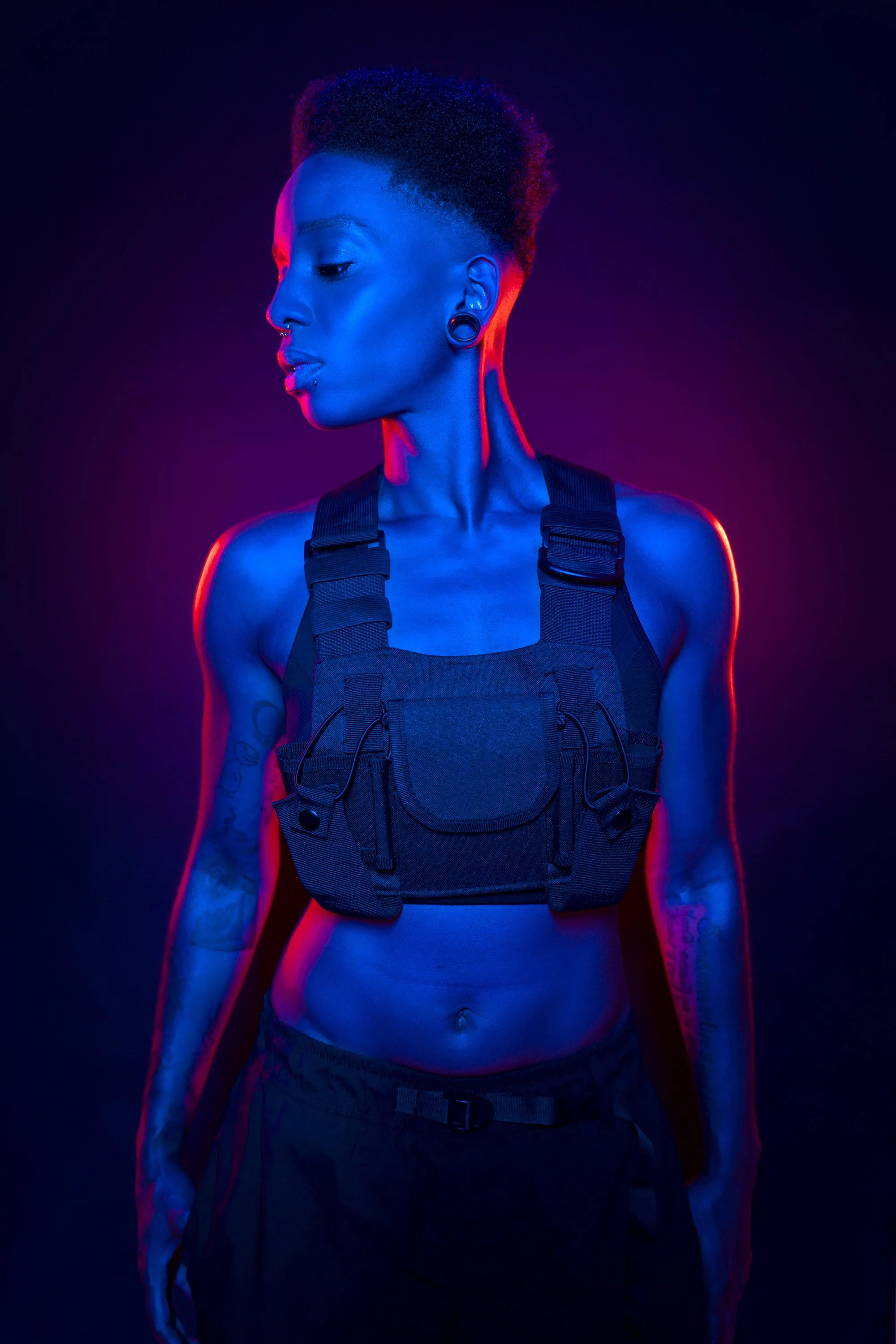 A young Black woman with short Afro hair wearing a chunky military style vest top. She is lit with blue light, with hard red edge lighting. stylised commercial portrait photography by Steve Bright