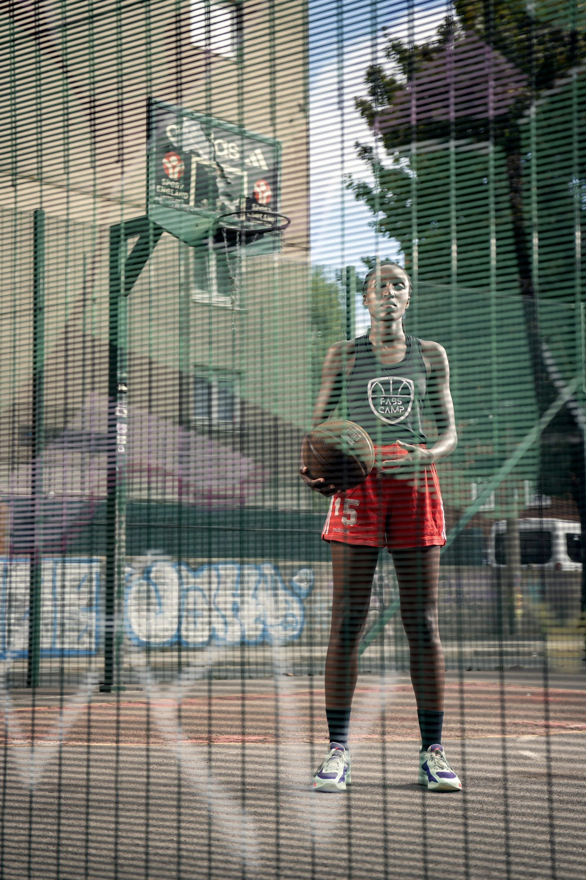A young woman standing on an outdoor basketball court, holding a basketball, viewed through a chain-link fence which has been sprayed with graffiti. dynamic sports  portrait photography by Steve Bright